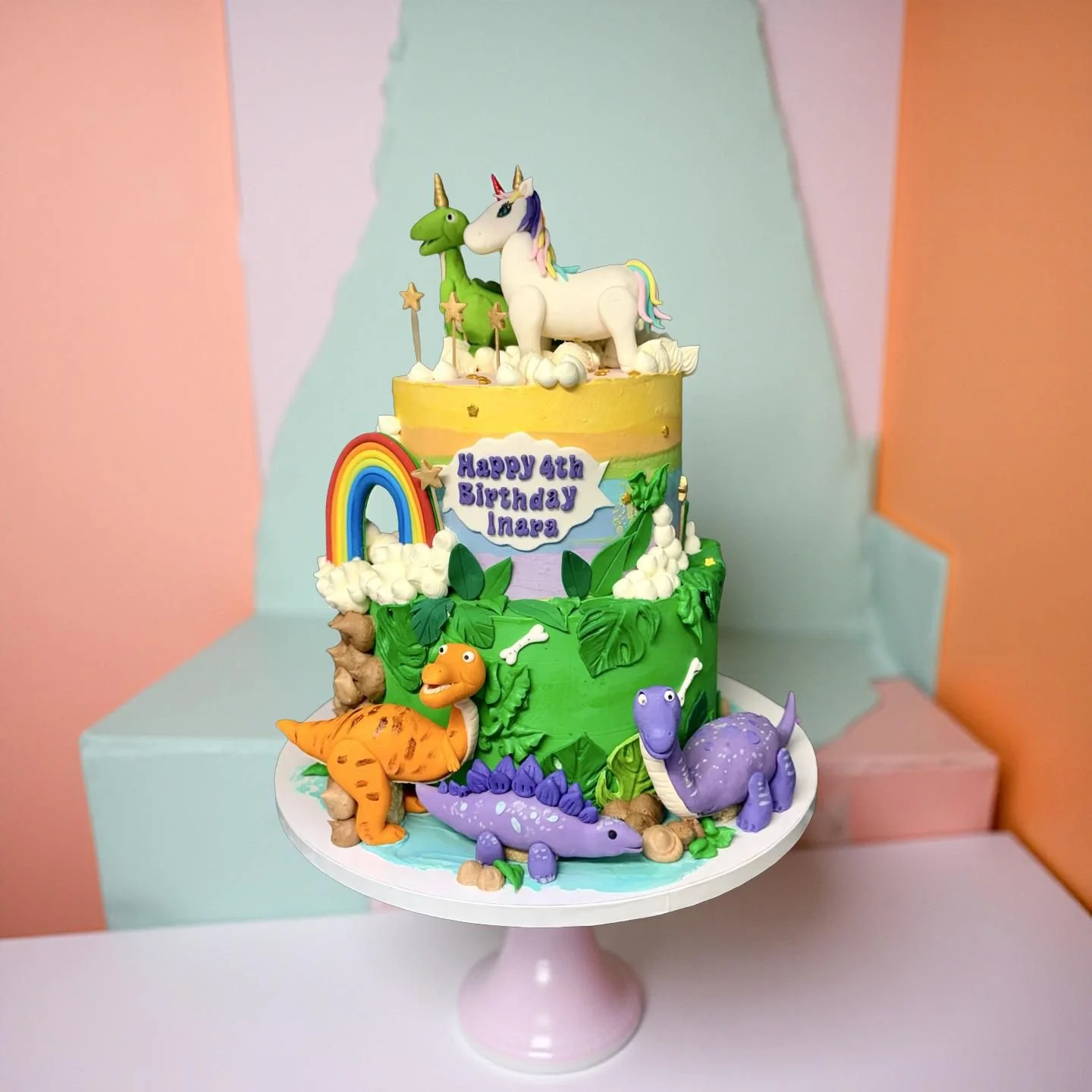 Colorful birthday cake with dinosaur and unicorn decorations, a rainbow, and a sign that reads 'Happy 4th Birthday Inara', set in a pastel-colored photo backdrop.
