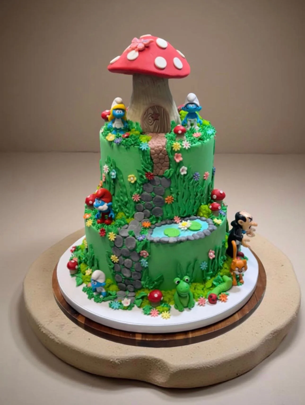 A colorful multi-tiered cake decorated with a mushroom house, Smurfs, and other characters from the Smurfs cartoon, with a pond, flowers, and greenery.