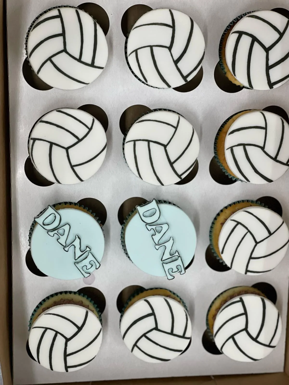 Cupcakes decorated with volleyball designs and one cupcake with 'DAD' written on top, arranged in a white gift box.