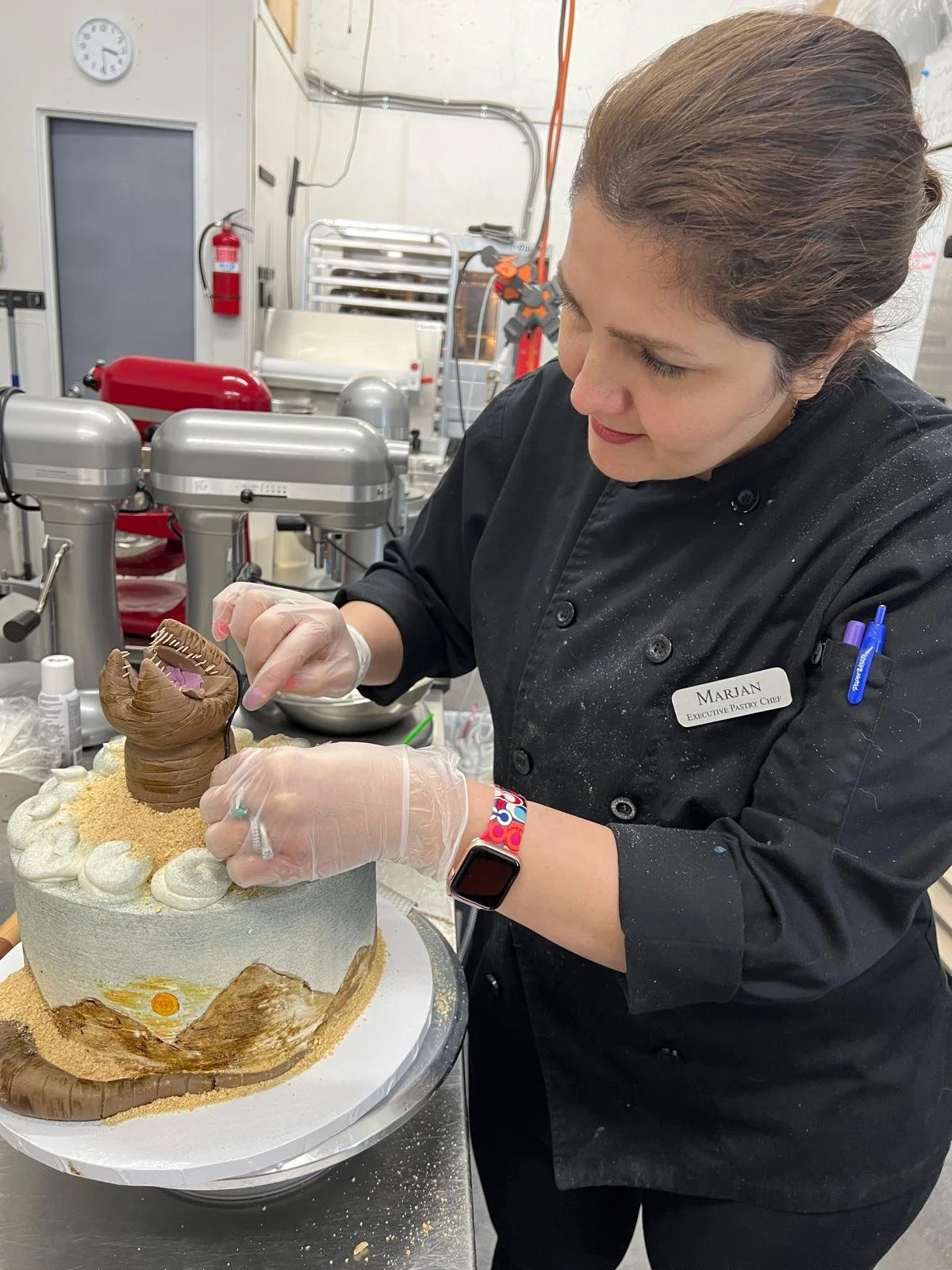 A woman in a black chef coat with a name tag that reads "Marjan" decorating a cake with a chocolate sculpture in a professional kitchen.