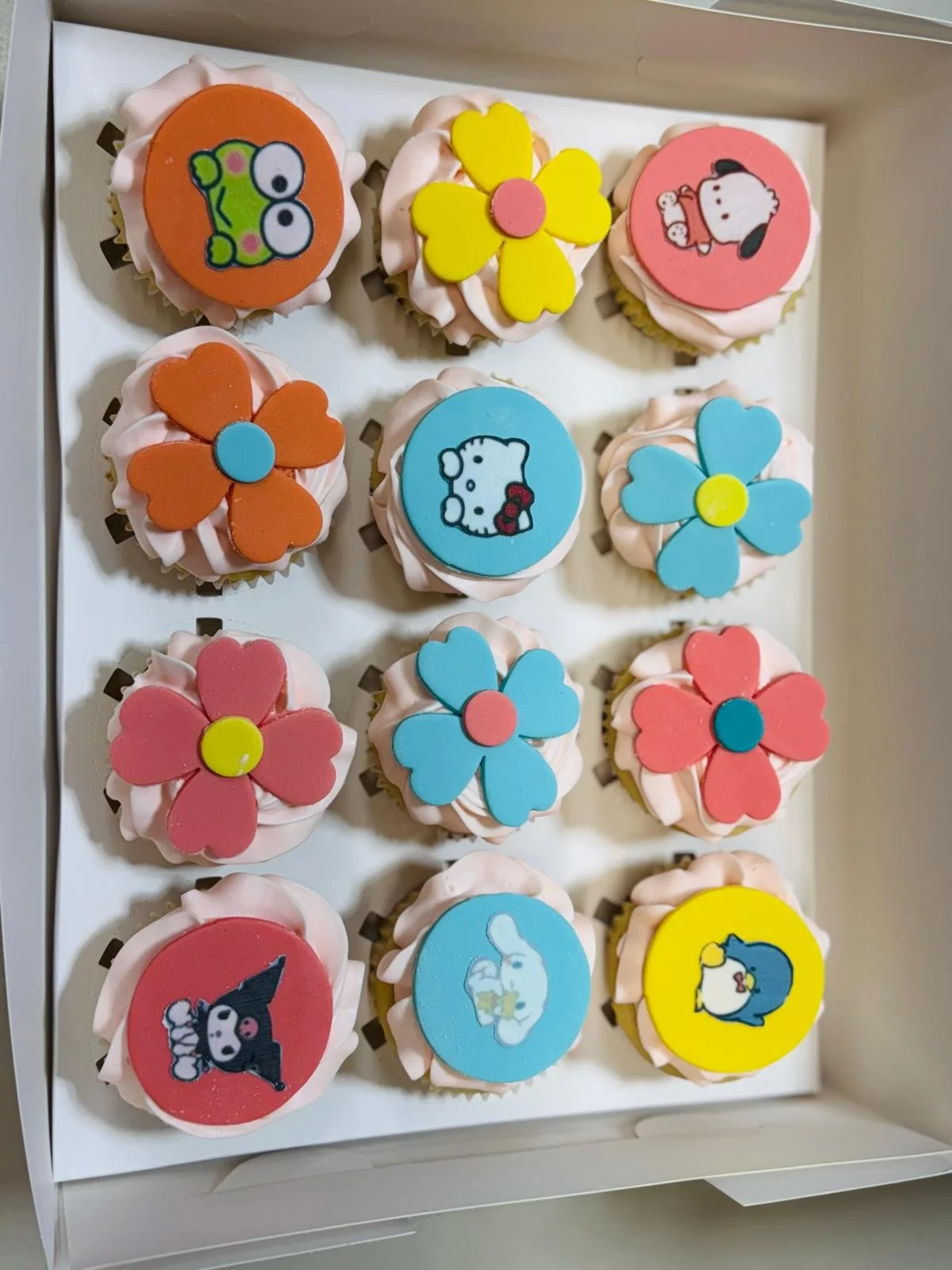 A box of decorative cupcakes with cartoon animal and flower toppers. The cupcakes have pink frosting and colorful fondant toppers including a frog, a dog, a cat, a bear, and flowers.