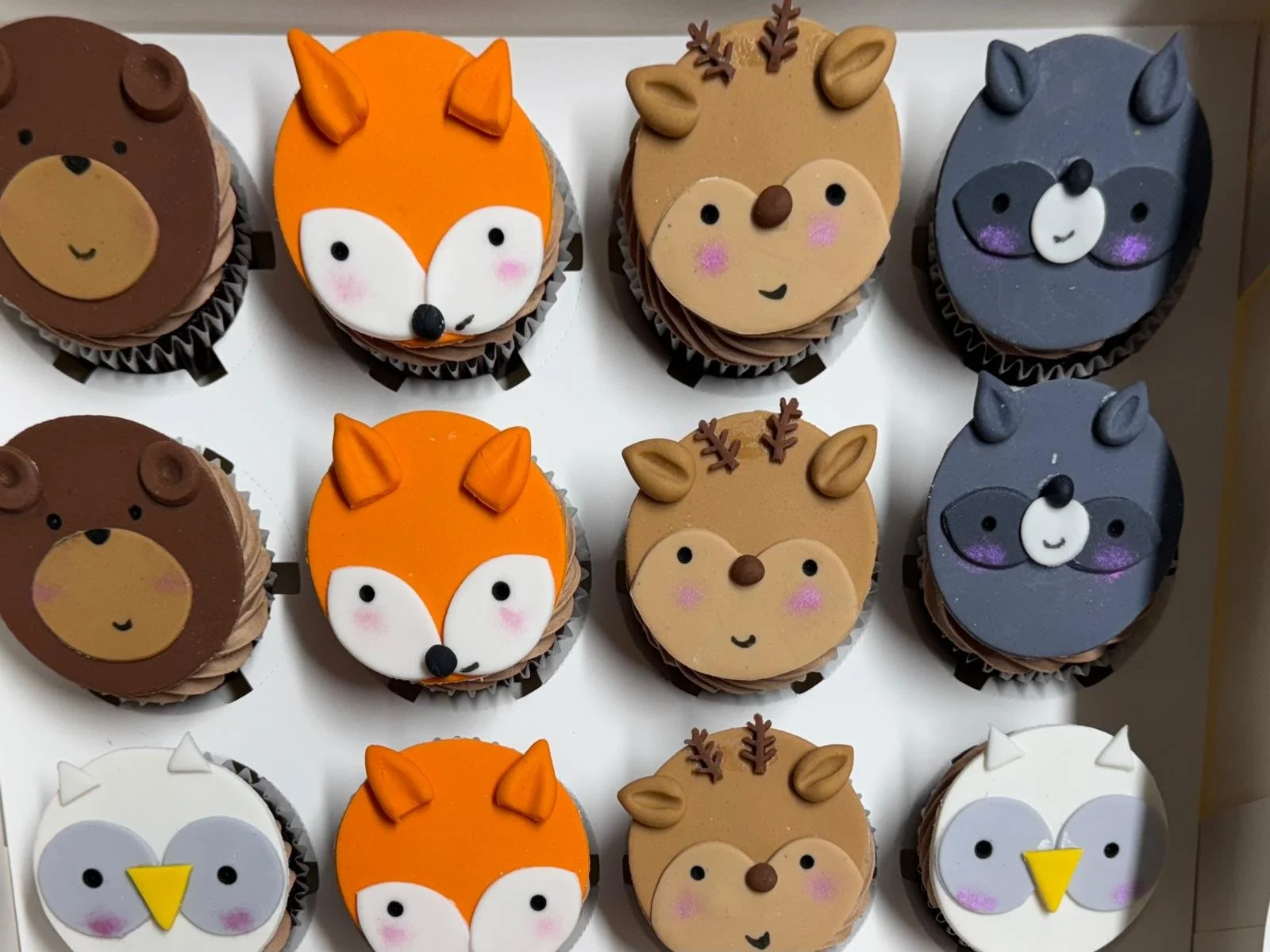 A tray of cupcakes decorated with animal faces, including bears, foxes, reindeers, wolves, penguins, and owls, each with colorful icing and cute facial features.