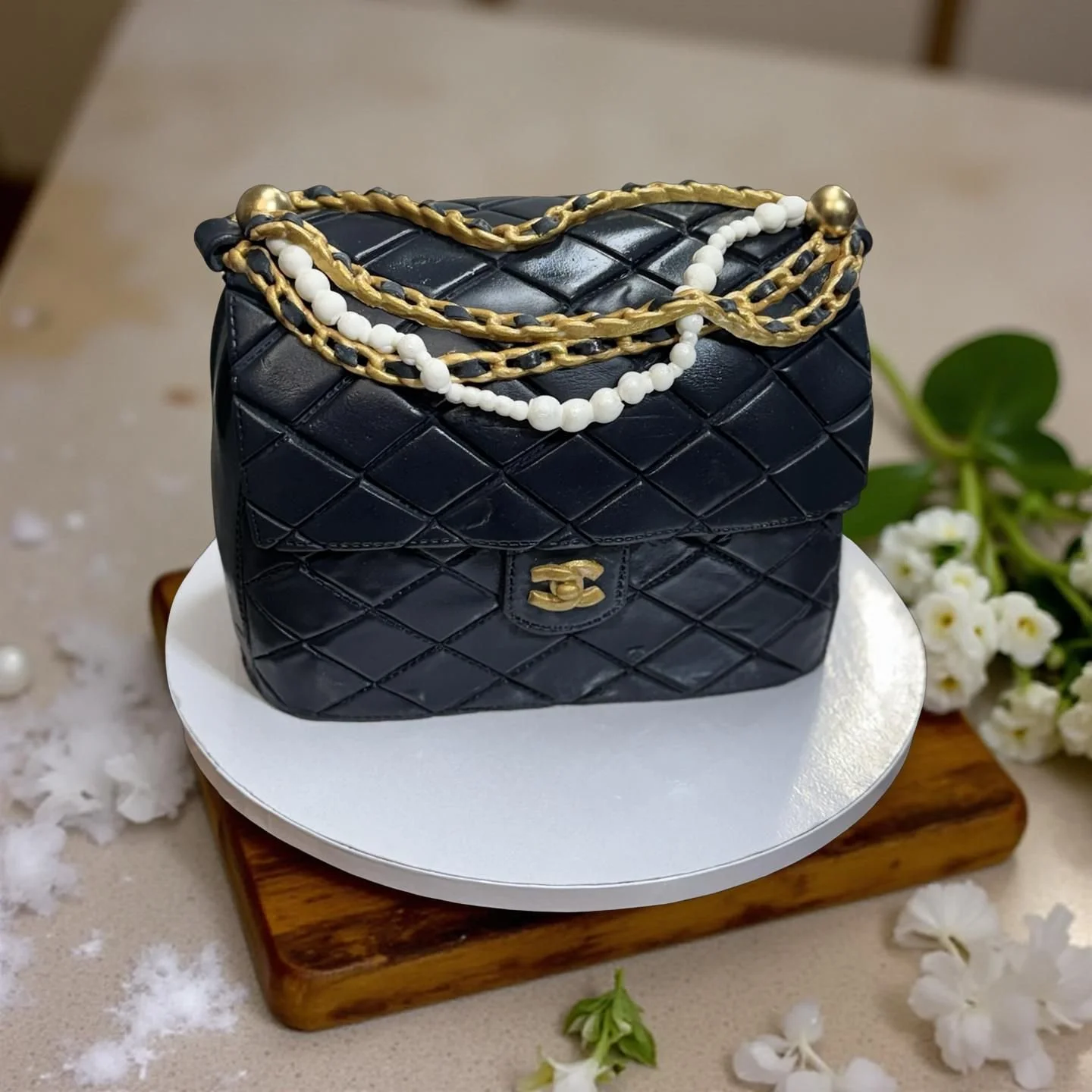A cake shaped like a black quilted designer handbag, decorated with gold and black chain details and a white pearl bead necklace, on a white cake board with floral decorations.