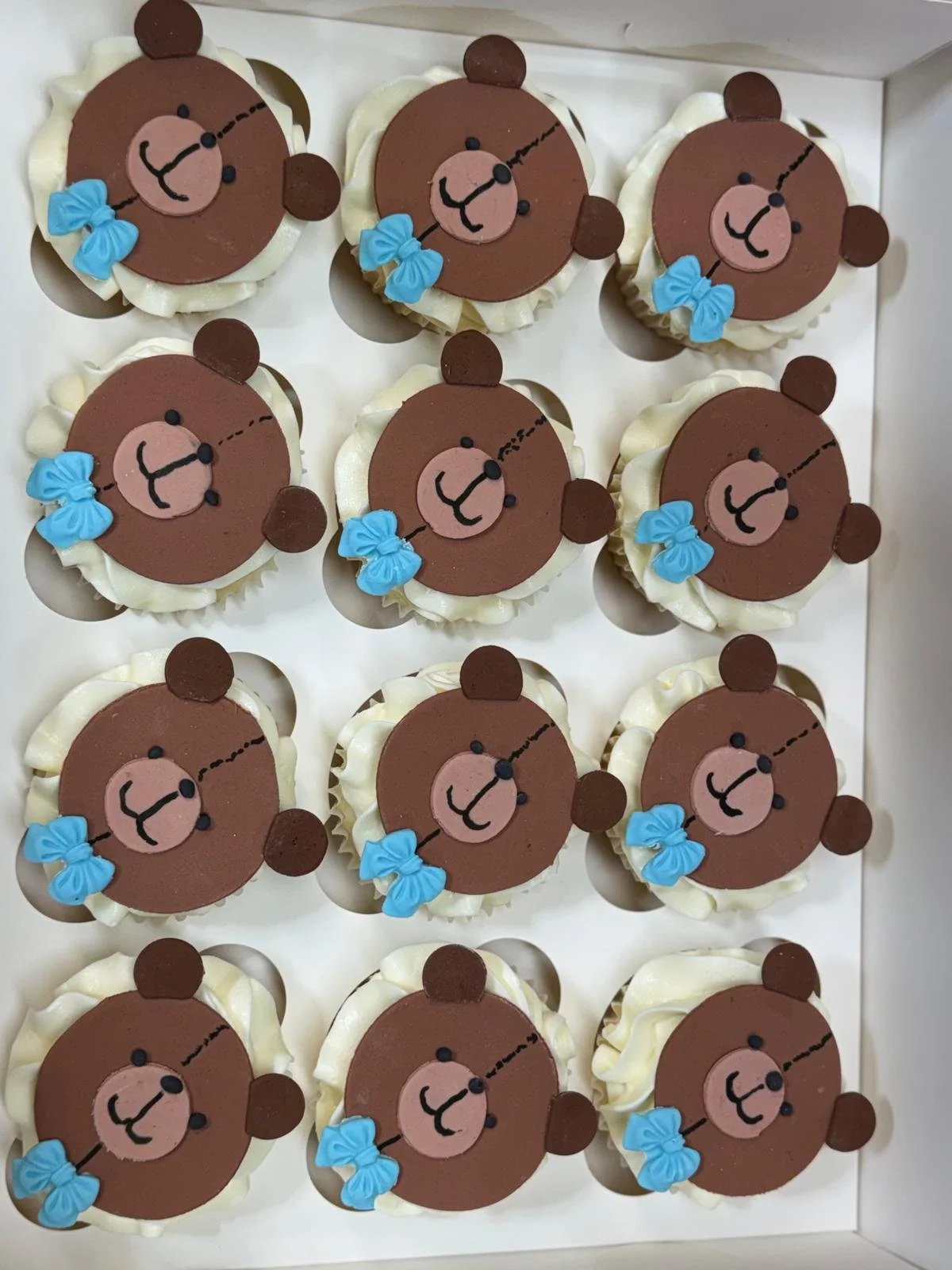 Cupcakes decorated with bear faces, featuring brown bear heads, light pink snouts, black eyes, a black nose, and a black line for a mouth, each with a blue bow.