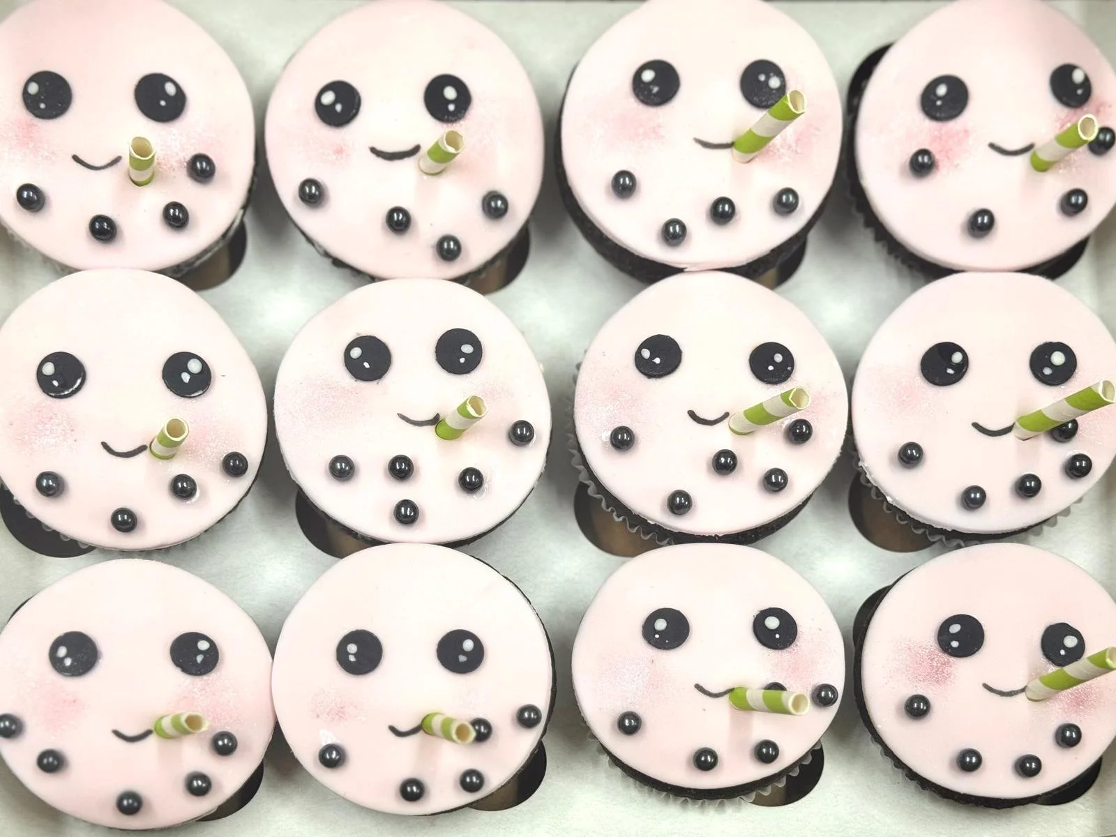 A tray of cupcakes decorated with pink and white icing, black eyes, and green-striped straws, resembling adorable faces.