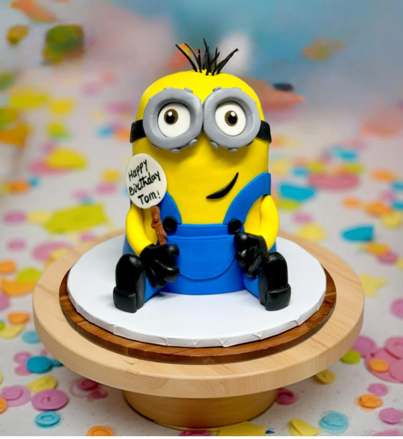 A Minion-themed birthday cake with yellow and blue fondant, featuring a Minion holding a small sign that says "Happy Birthday Tom!" with colorful confetti scattered on the background.