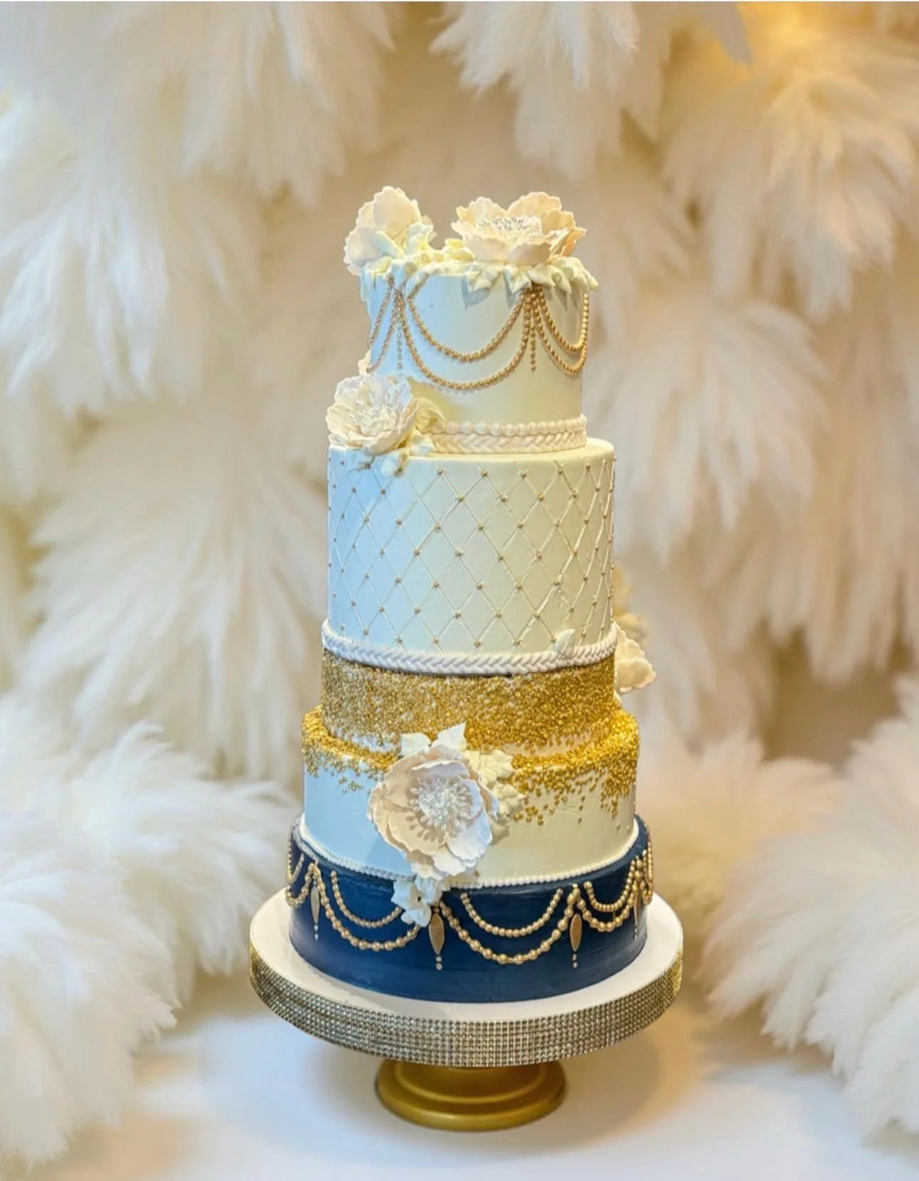 A tall, multi-tiered wedding cake decorated with white, gold, and navy blue elements, including floral decorations, pearls, and chains, set against a soft, feathered background.