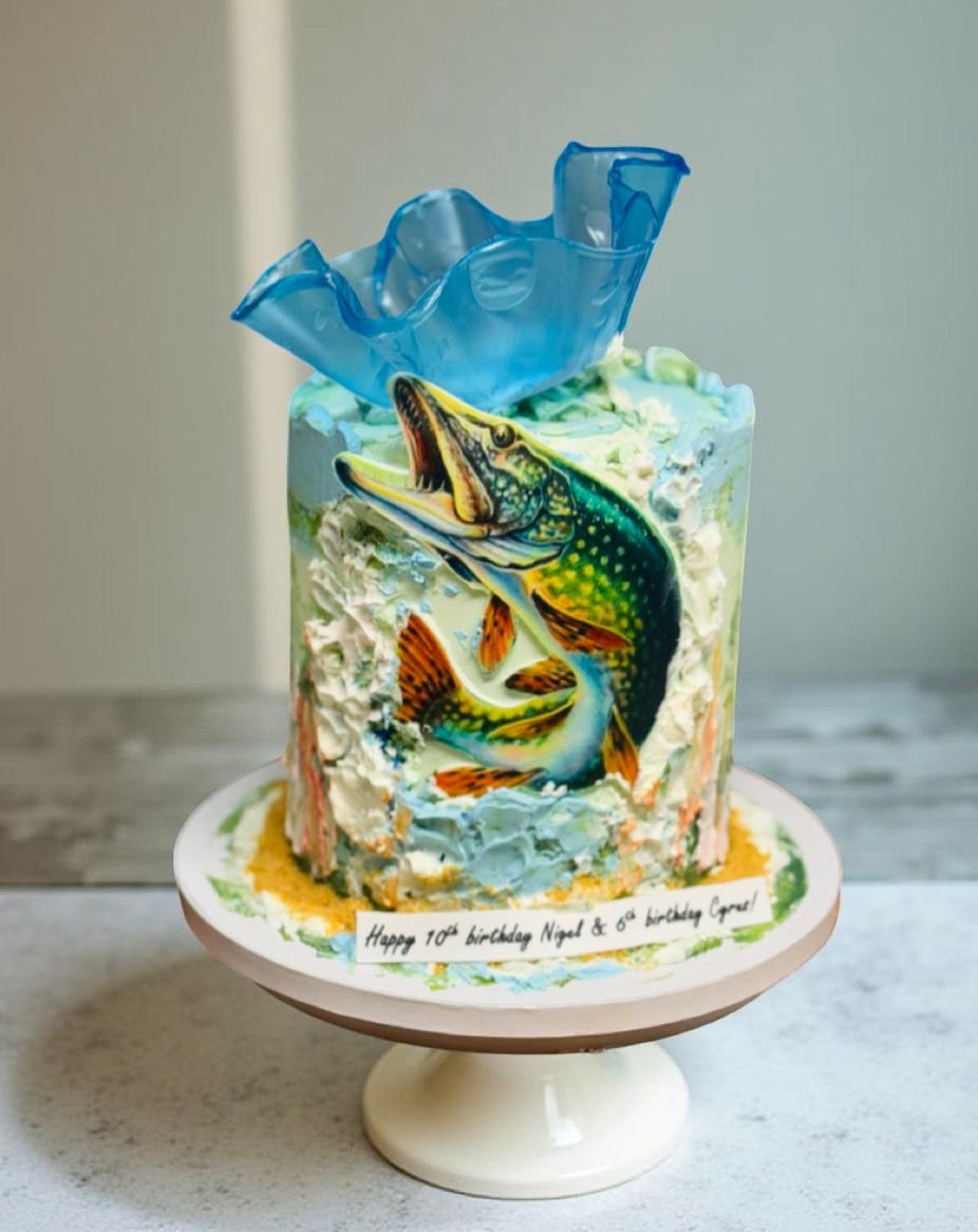 A birthday cake with a colorful fish and water splash decoration on top, featuring a large fish leaping out of water, with a message on the cake reading 'Happy 10th birthday Nigel & 6th birthday Lara!'