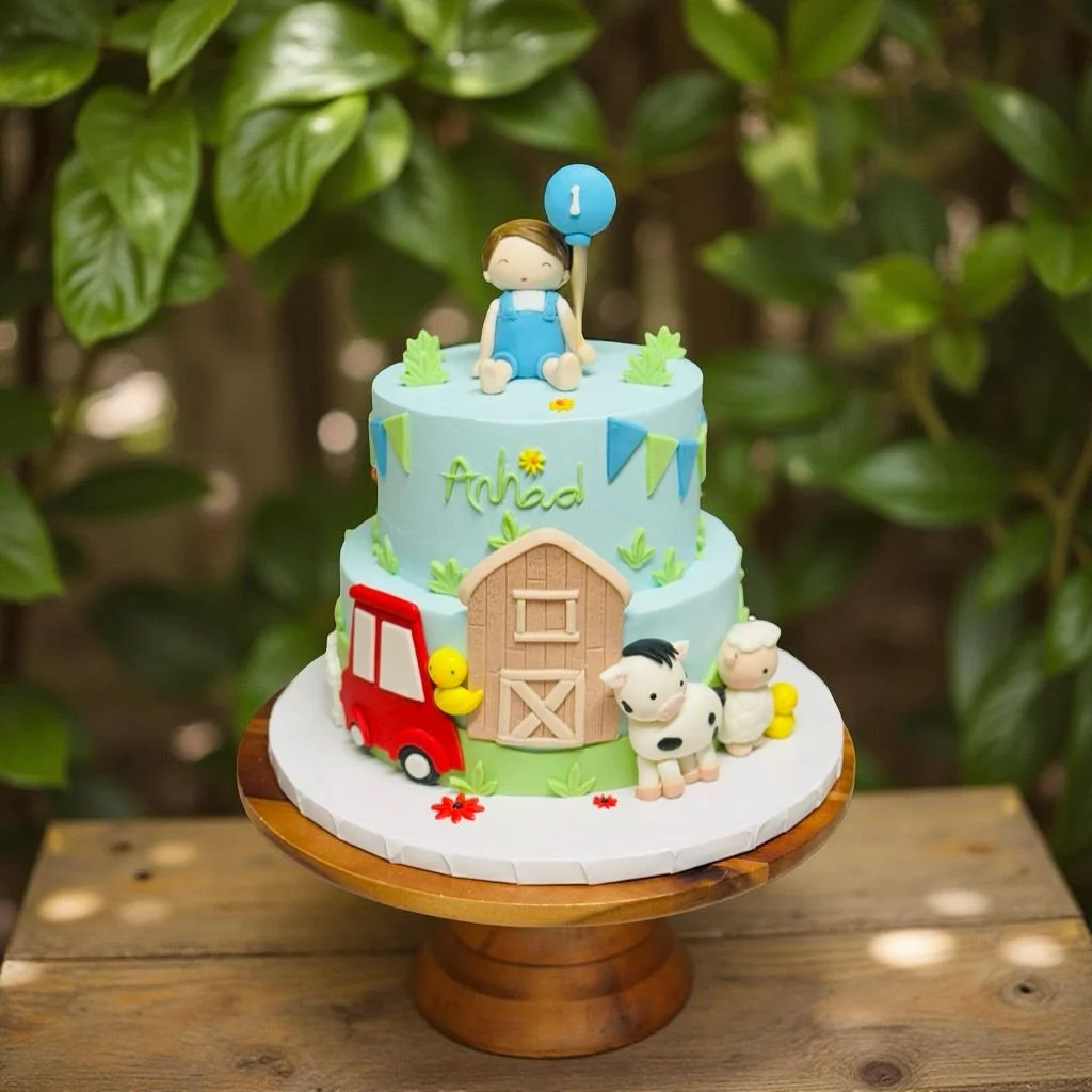 A two-tiered birthday cake with farm-themed decorations, featuring a figurine of a boy sitting on top holding a balloon with the number 1. The cake has fondant animals, a barn, a red tractor, and green foliage. The name 'Anibal' is written on the cak