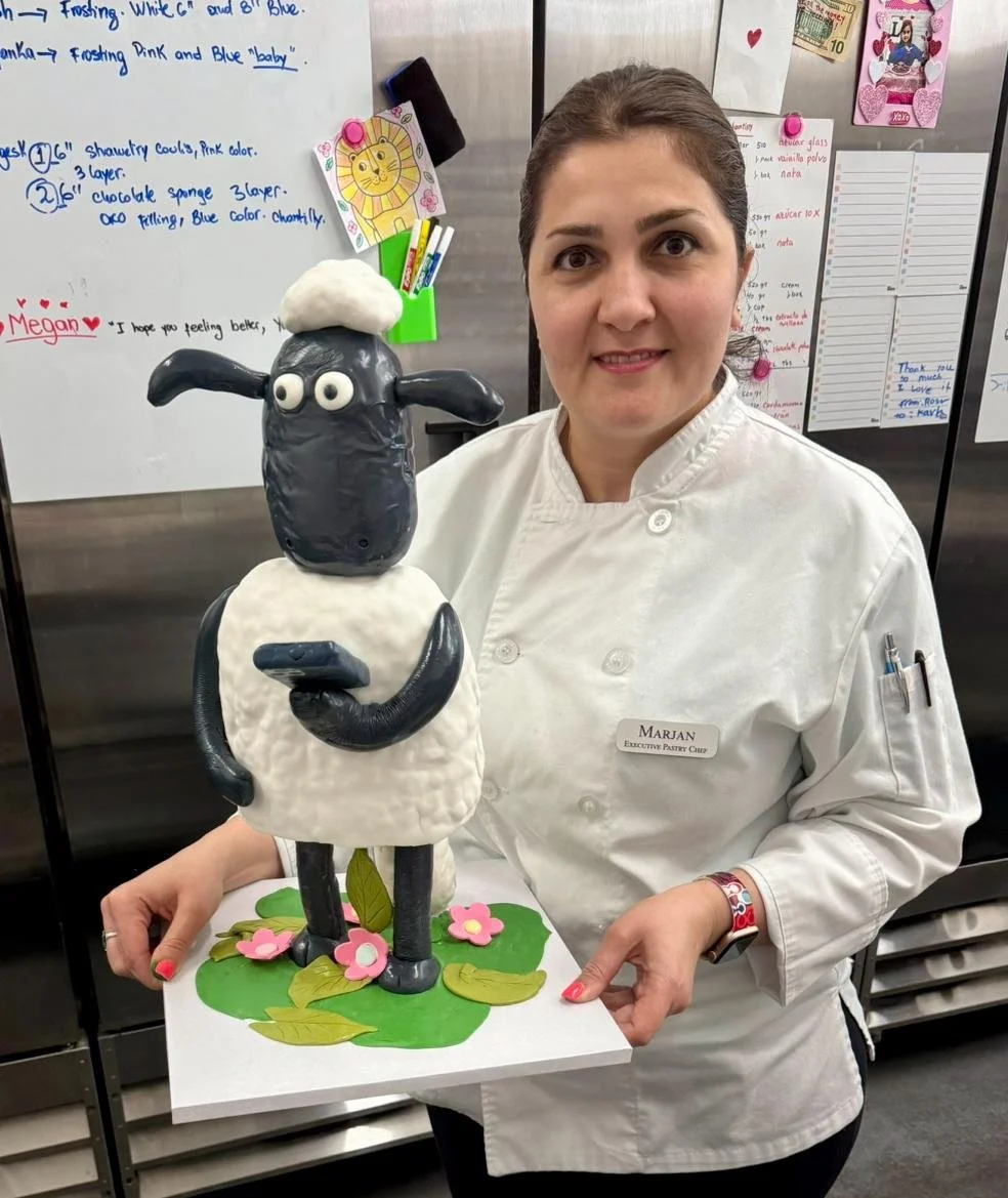 A woman in a white chef uniform holding a decorative cake sculpture of Shaun the Sheep with a sheep's head on top, standing on a green base with pink flowers. The woman is smiling in a kitchen or bakery setting.