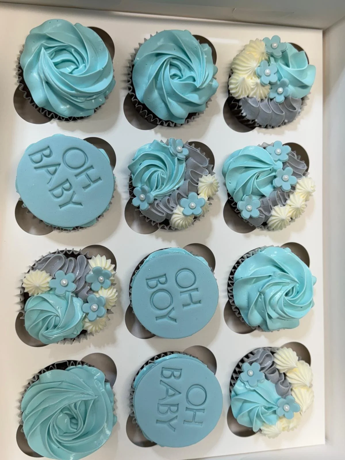 Box of decorated cupcakes with blue, gray, and white frosting, some with blue flower decorations and the words "OH BABY" written on some of the blue fondant discs.