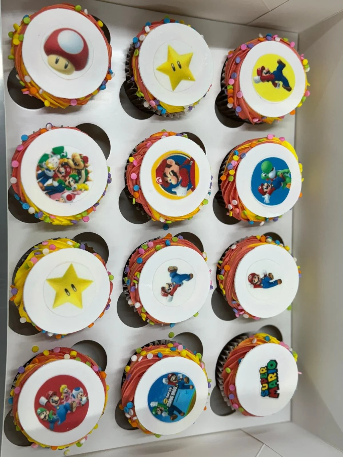 A collection of nine colorful cupcakes decorated with Super Mario themed edible images, frosting, and rainbow sprinkles, arranged in a white cupcake holder.