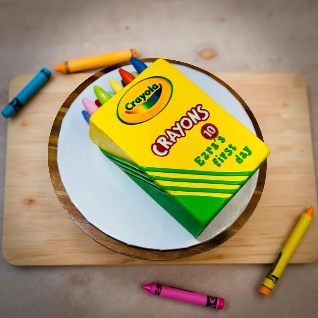 A cake designed to look like a box of Crayola crayons on a wooden table, with colorful crayon decorations around it.