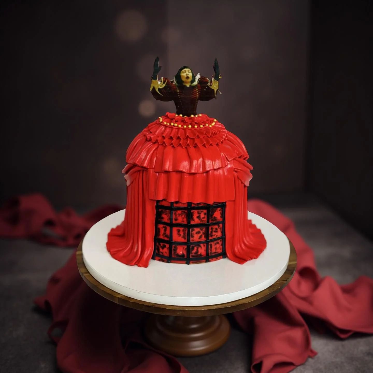 A cake designed to look like a woman's dress, with a red ruffled skirt, on a white cake board. The top of the cake features a figure of a woman with short black hair, dressed in a dark red and black outfit, with her arms raised.