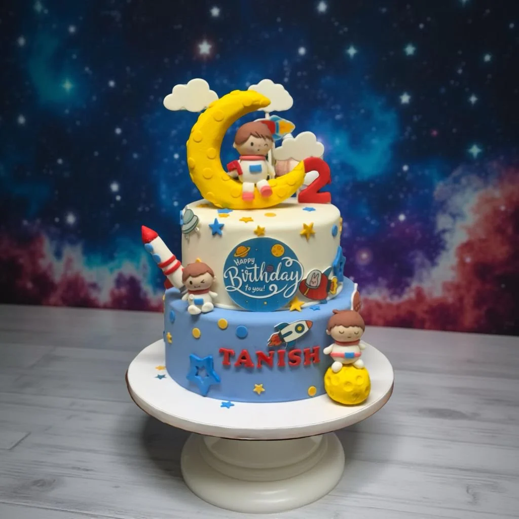 Space-themed birthday cake with astronaut and moon decorations, featuring the name Tanish in red letters and a "Happy Birthday" message, with a cosmic background.