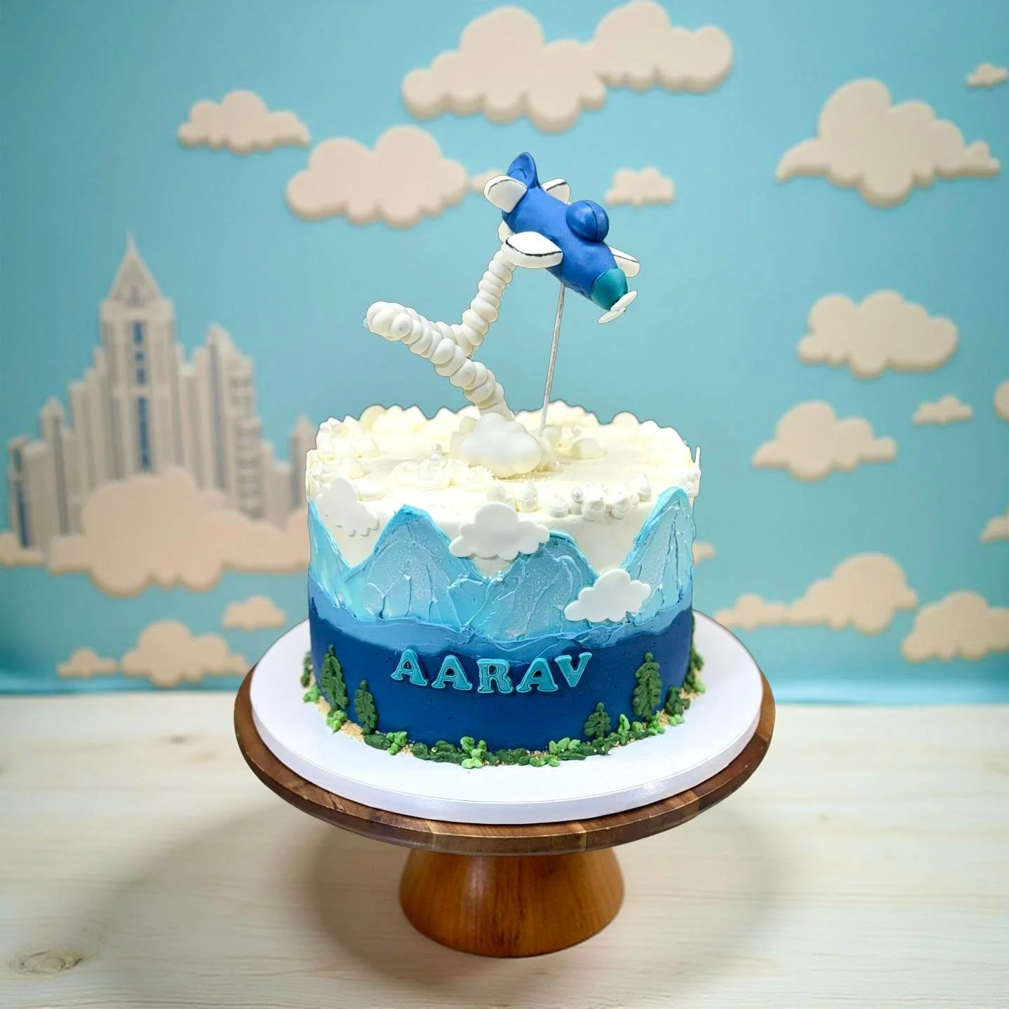 A cake decorated with mountain and cloud designs, featuring the name 'AARAV' on the front, topped with a blue and white airplane made of fondant or similar material, set against a sky-themed backdrop with clouds and cityscape.