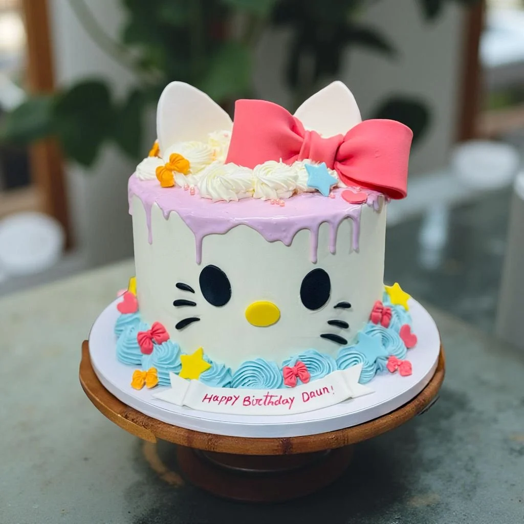 Hello Kitty themed birthday cake decorated with pink, blue, yellow, and red frosting, a large pink bow, white ears, and colorful stars and bows, with a message that reads "Happy Birthday Daun!"