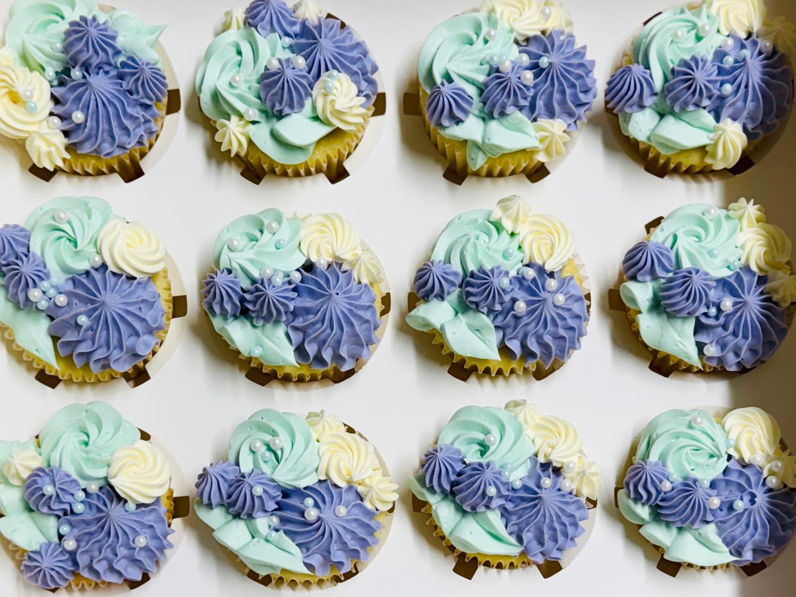 A tray of twelve cupcakes decorated with pastel blue, purple, and white frosting with pearl sprinkles.