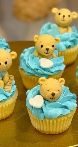 Cupcakes decorated with blue frosting and bear-shaped fondant toppers, some with closed eyes and smiling, others with open eyes.