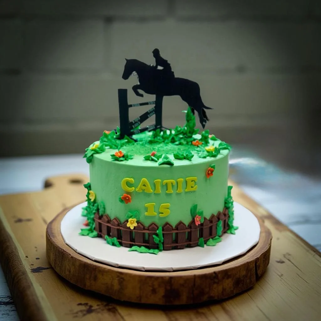 A birthday cake with green icing, decorated with small colorful flowers, featuring a black silhouette of a person riding a horse on top, celebrating Caitie's 15th birthday.