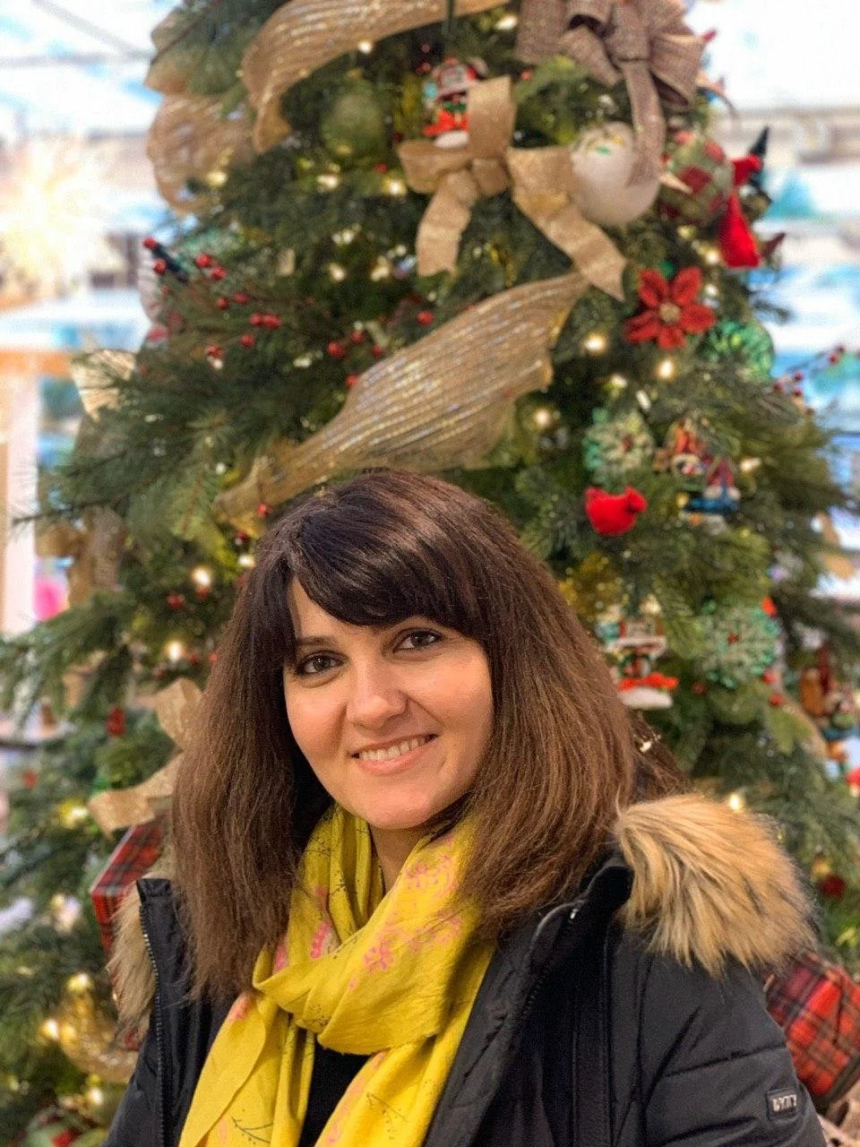A woman with shoulder-length brown hair, wearing a yellow scarf and black coat, standing in front of a decorated Christmas tree with ornaments, ribbons, and lights.