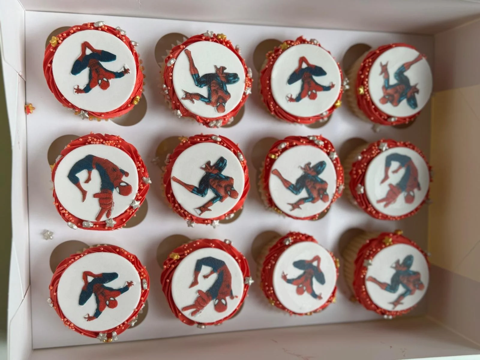 A box of cupcakes decorated with edible images of Spider-Man in various poses, with red icing and pearl and star-shaped sprinkles.