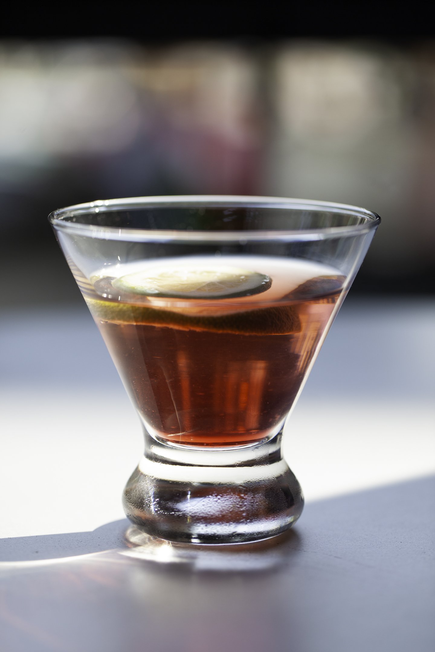 A glass filled with a red cocktail garnished with a lime slice, placed on a reflective surface with a blurred background.