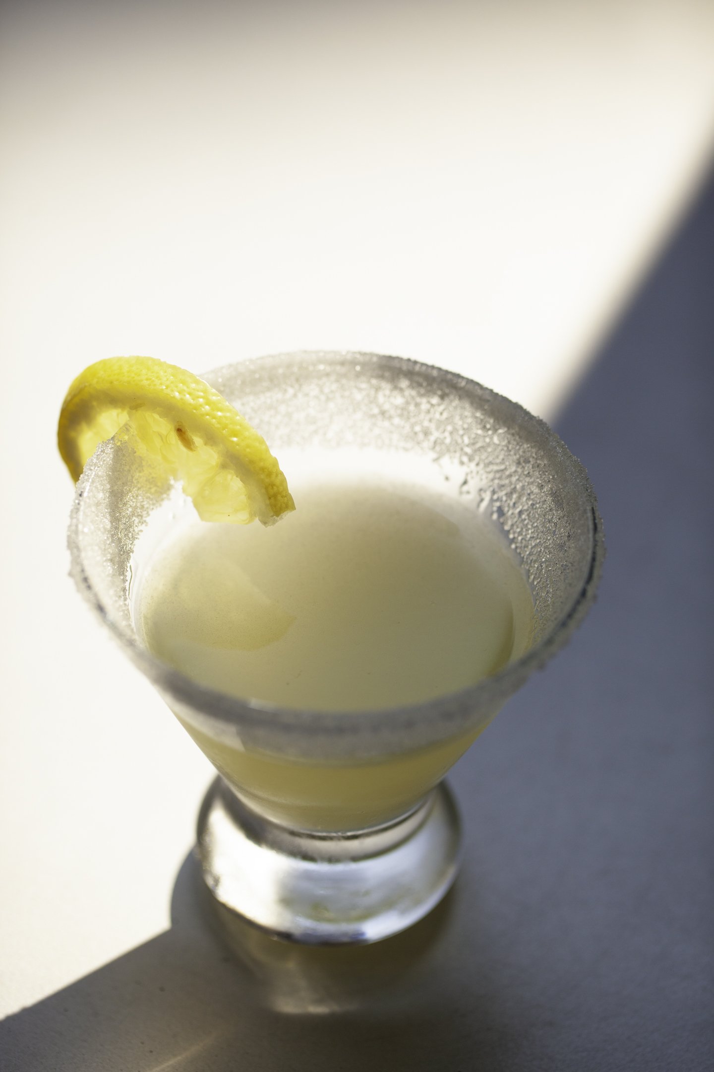 A close-up of a margarita cocktail in a glass with a salted rim, garnished with a lemon wedge, sitting on a white surface with shadows.