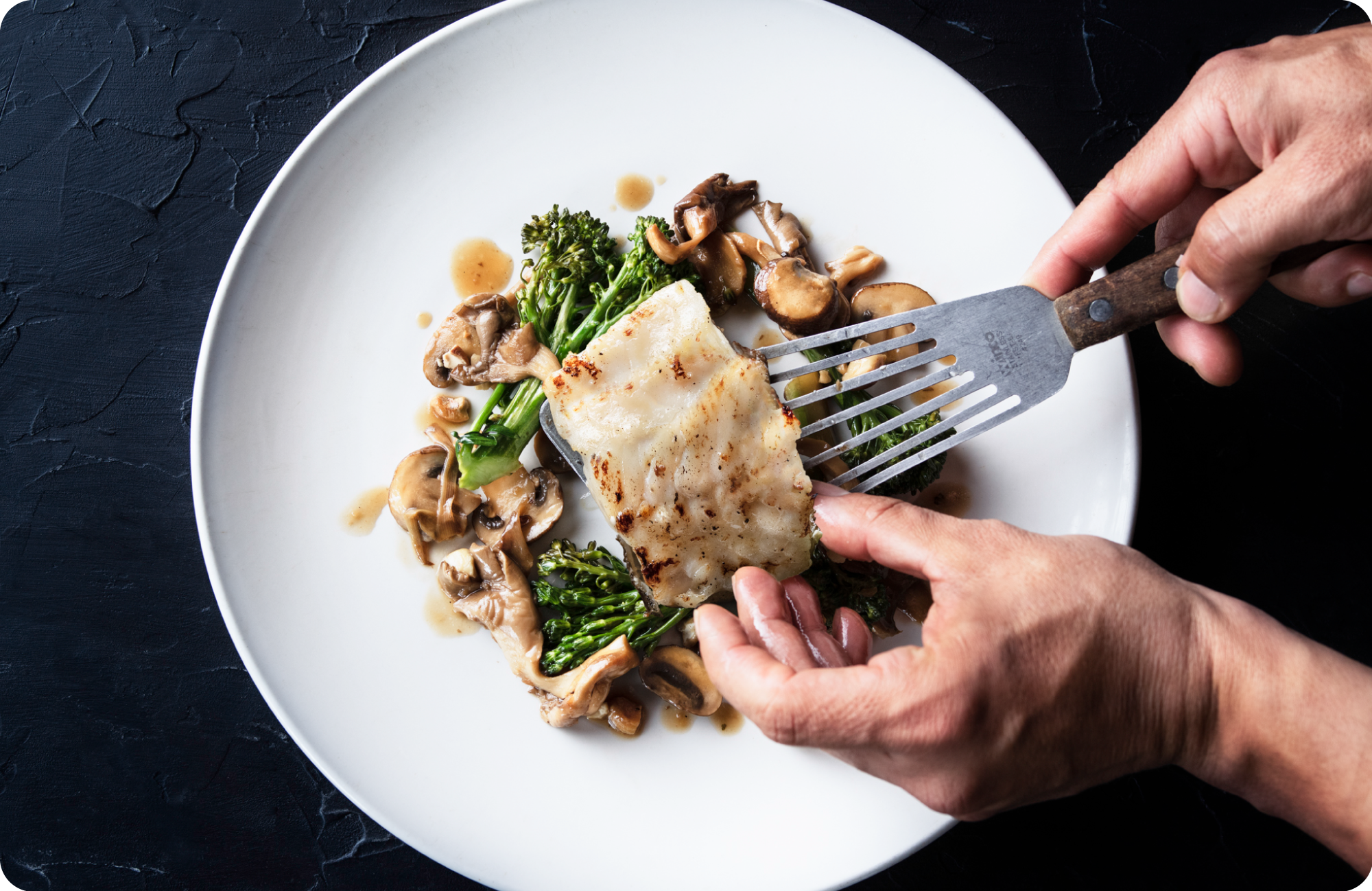 Person holding fork with a piece of baked fish, served with sautéed mushrooms and broccoli on a white plate, set on a dark surface.