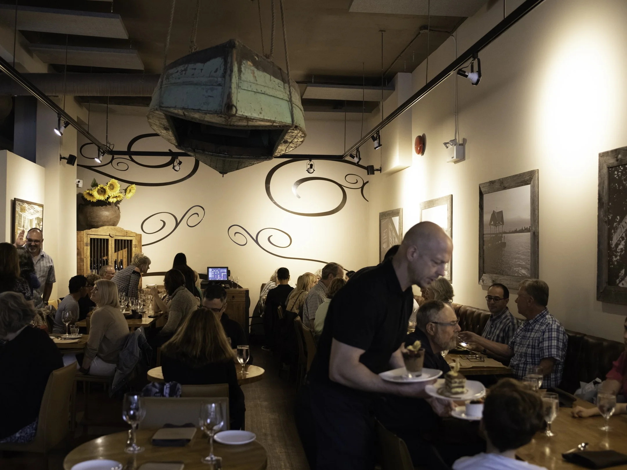 A crowded restaurant with many people seated at tables, some eating and some engaged in conversation. A waiter in the foreground is serving dessert. A large, vintage, boat-shaped light fixture hangs from the ceiling. The walls are decorated with framed pictures and a sunflower arrangement in a vase.