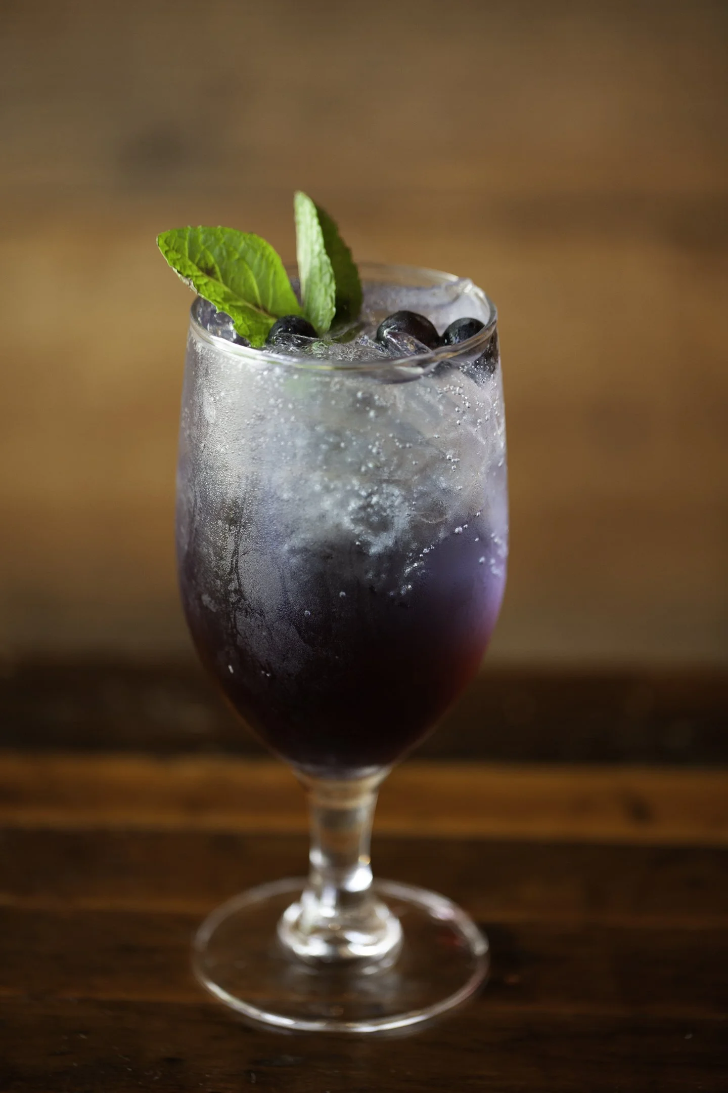 A tall glass filled with a dark purple or black beverage, ice, garnished with fresh mint leaves and blueberries, sitting on a wooden surface with a warm, blurred background.