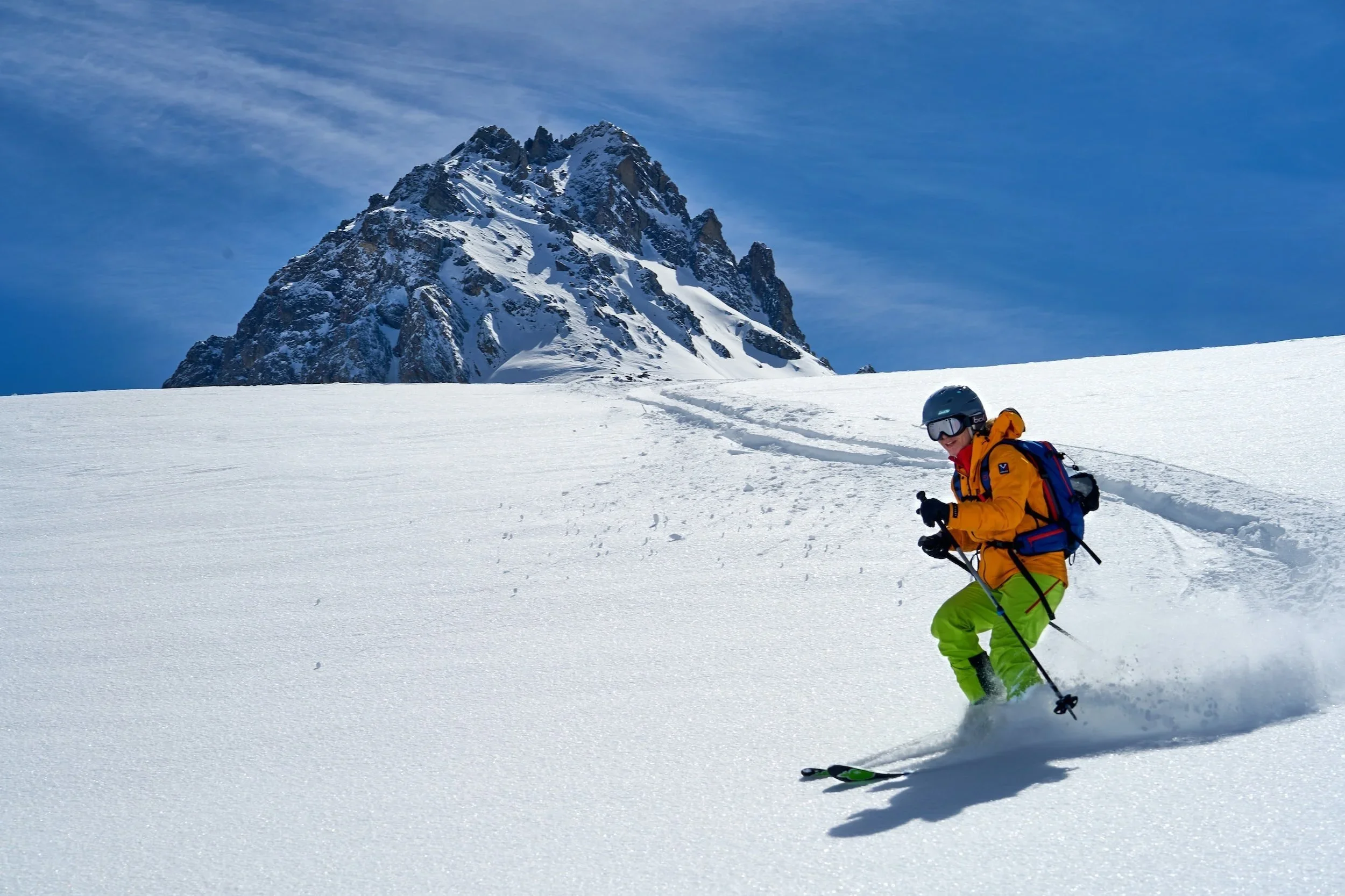 Skier in a bright orange jacket and lime green trousers carving confidently through deep snow on a wide mountain slope, representing the lasting movement confidence The Movement Bank™ programmes are designed to build.