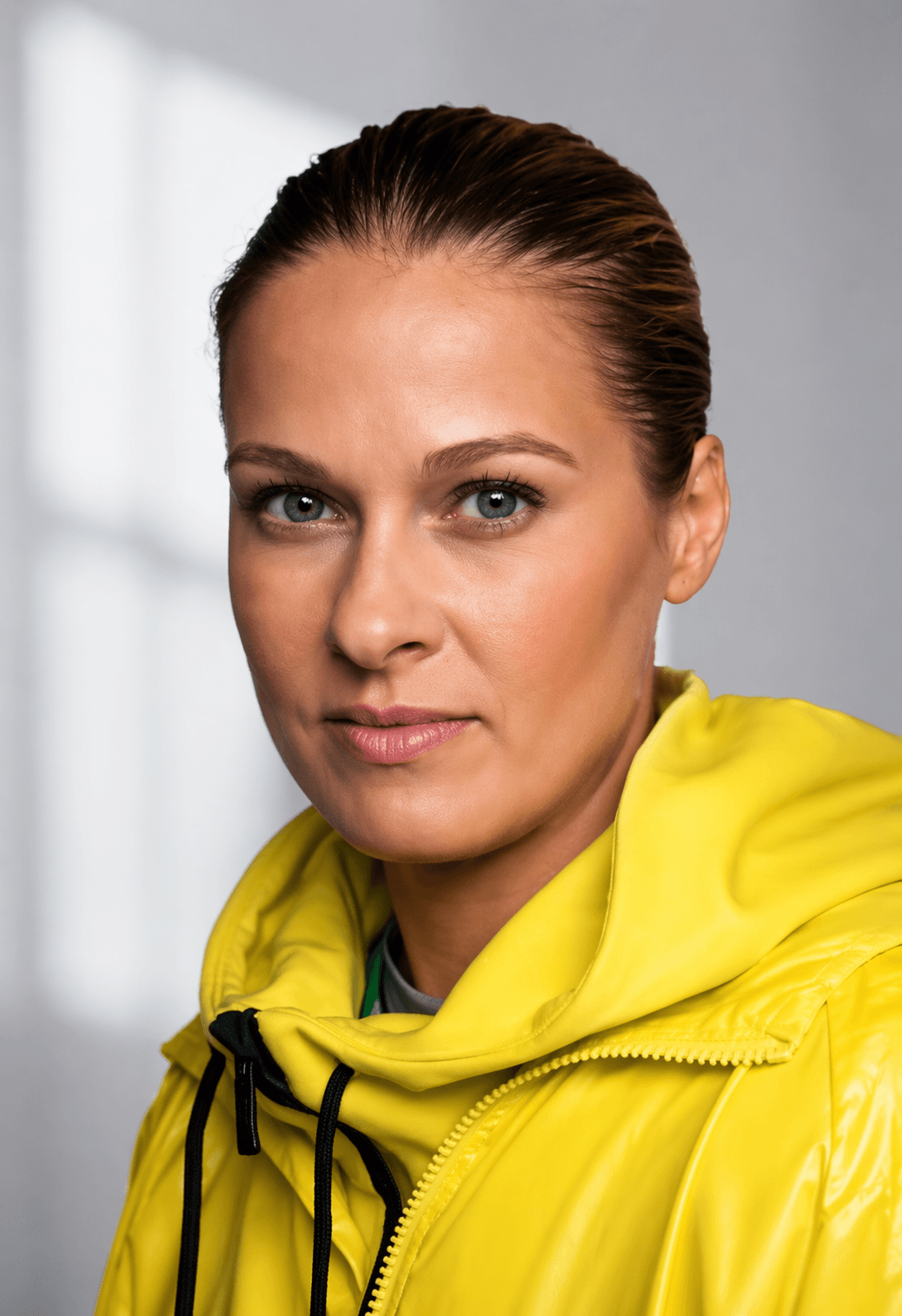 Close-up headshot of Elizabeth Banks with her hair pulled back, wearing a bright yellow hooded jacket, looking directly at the camera with a composed and assured expression.