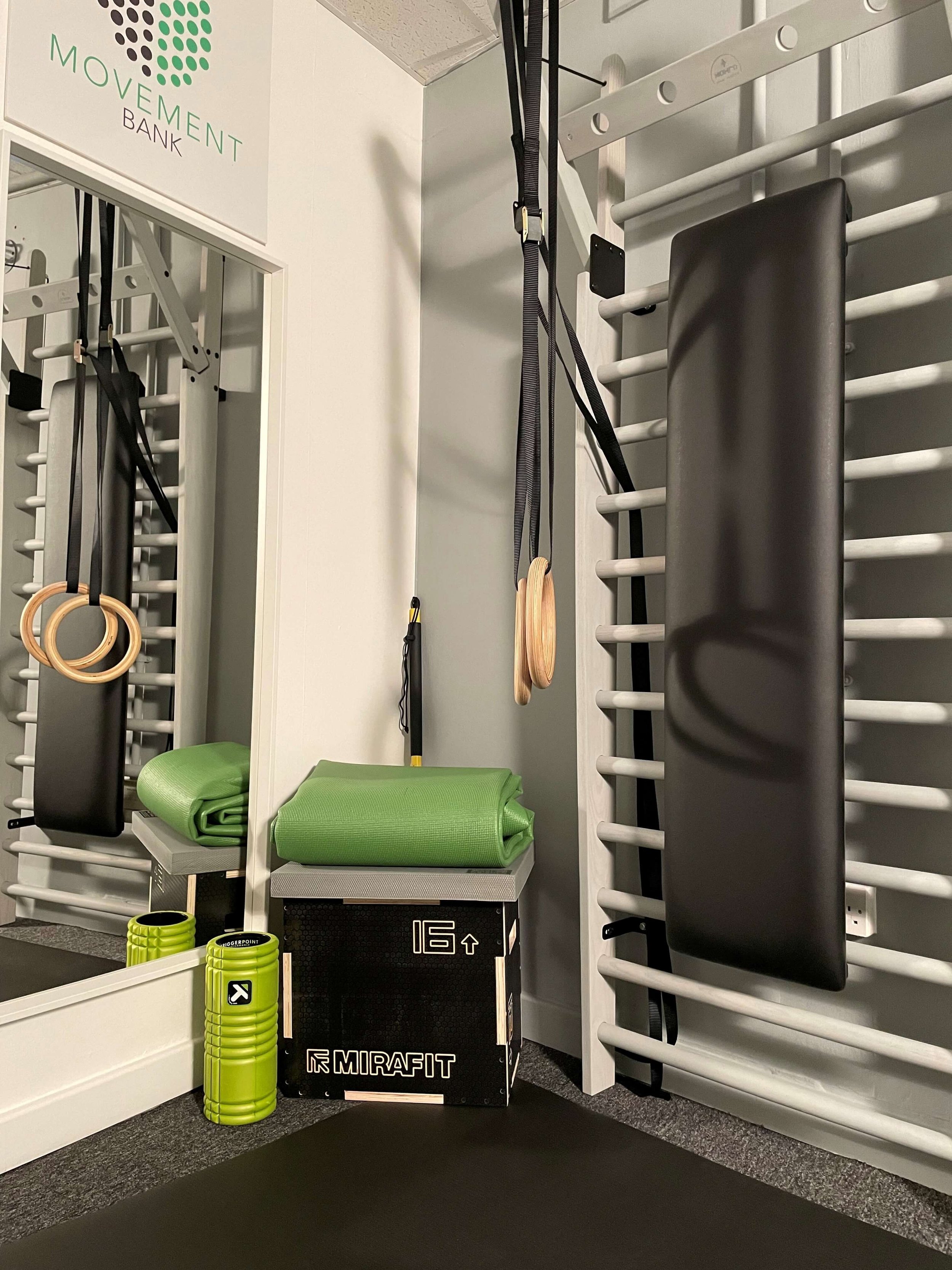 Calm, intentionally organised corner of The Movement Bank™ private studio in Liverpool Street, featuring wall bars, gymnastic rings, a plyo box with folded yoga mats, and specialist equipment arranged to support focused neurological work