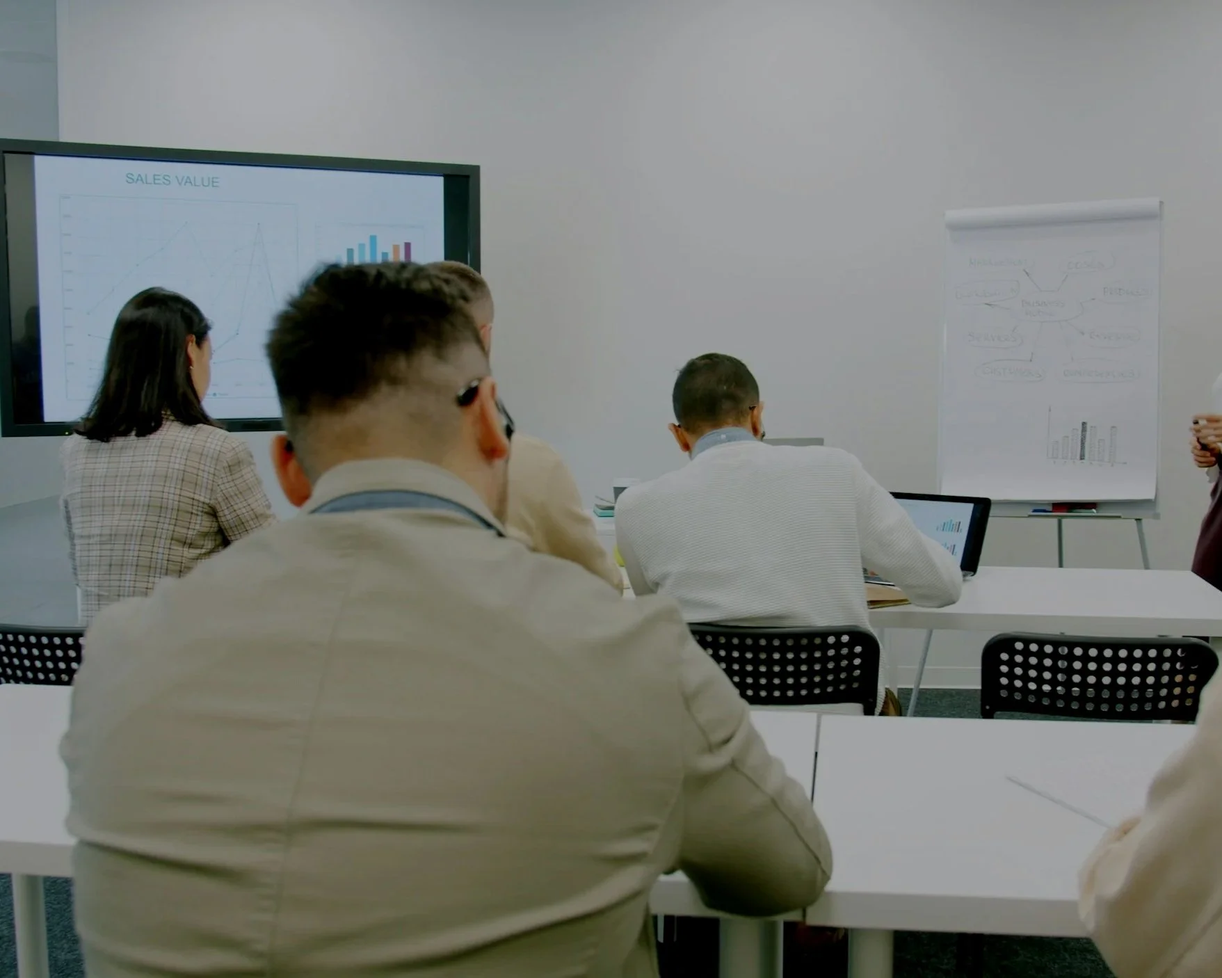 People attending a business presentation, sitting at a long table and watching a graph on a screen in a conference room.