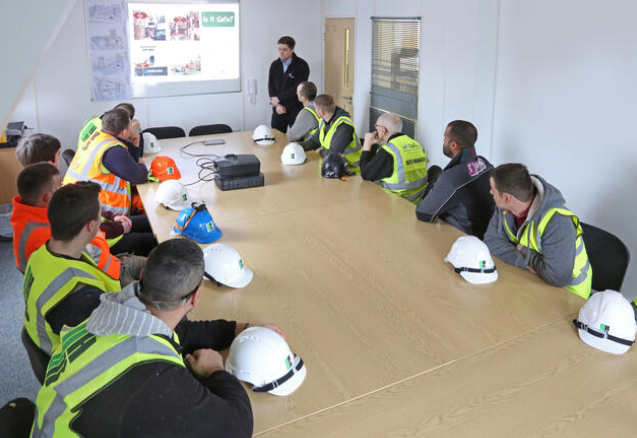 Group of construction workers attending safety training in a conference room, wearing safety vests and helmets, listening to instructor.