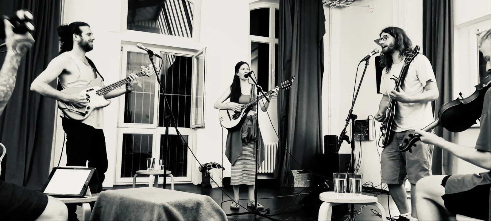 Eine Gruppe von vier Musikern spielt in einem Raum mit offenen Fenstern, gefüllt mit Instrumenten wie Gitarren und einem Mikrofon.