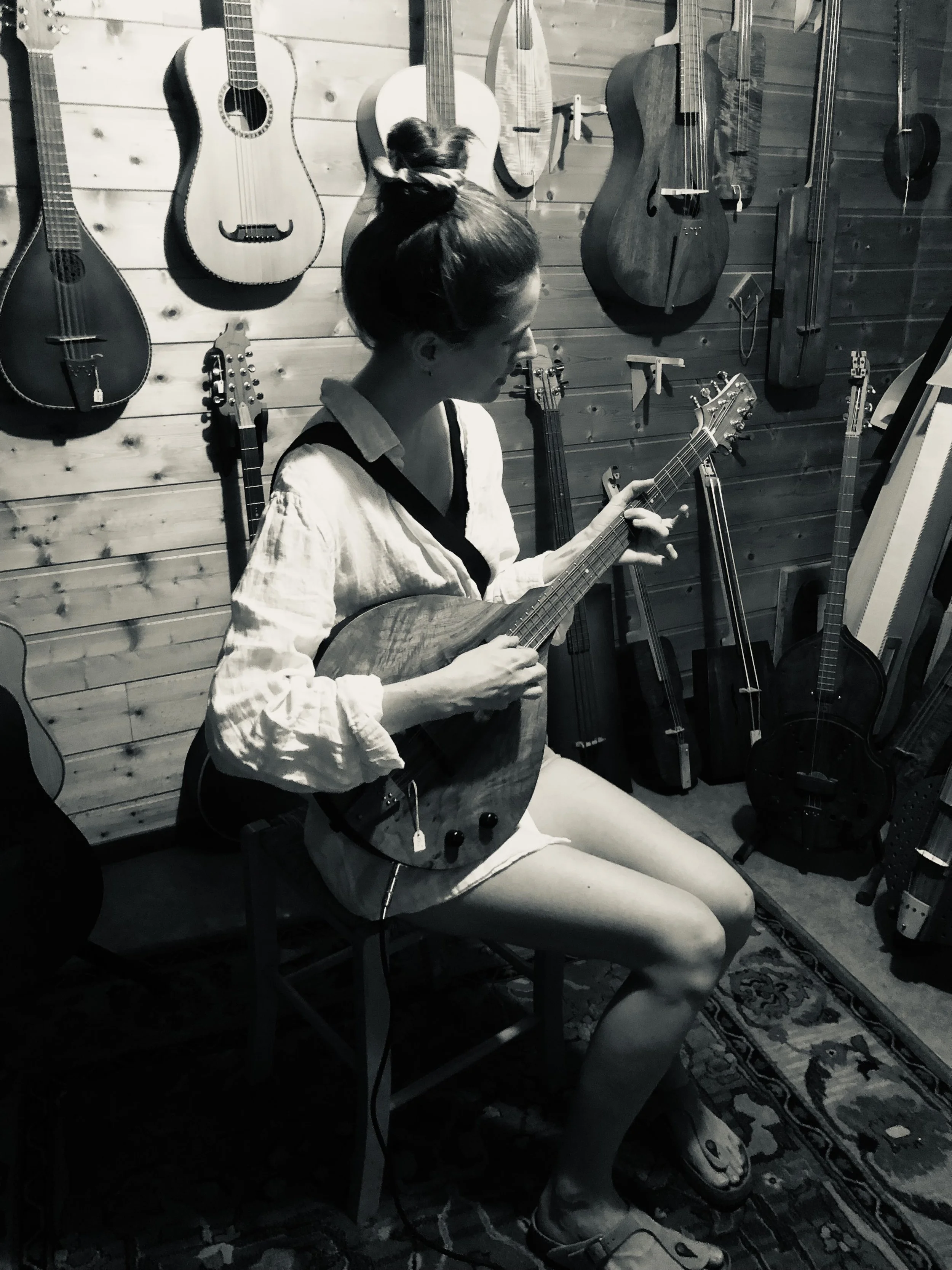 Eine junge Frau sitzt auf einem Stuhl in einem Musikgeschäft, spielt auf einer akustischen Irish Bouzouki. Der Hintergrund ist mit verschiedenen Saiteninstrumenten an einer Holzwand geschmückt.