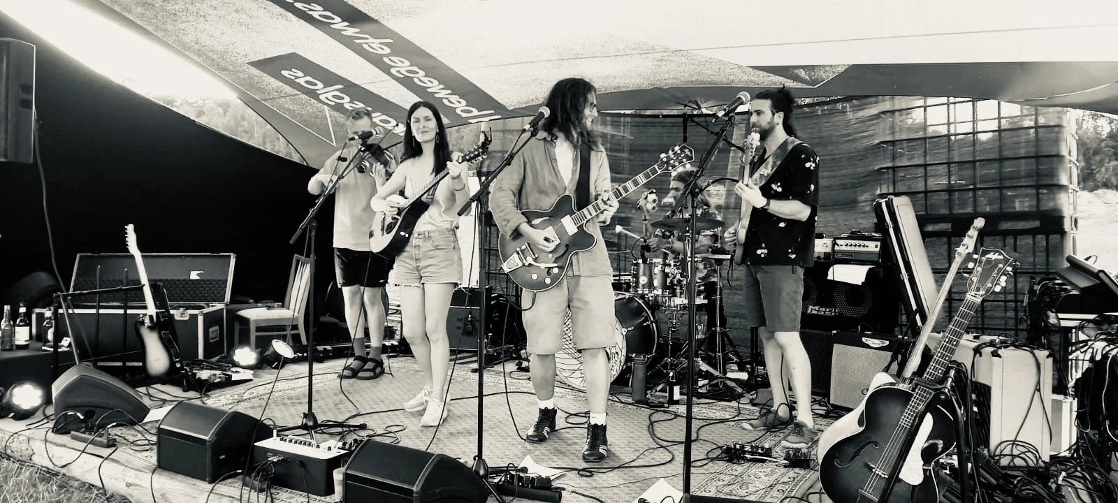 Eine Band spielt auf einer Bühne im Freien, unter einem Zeltdach. Die Band besteht aus fünf Mitgliedern mit Musikinstrumenten, darunter Saiteninstrumente und Schlagzeug.
