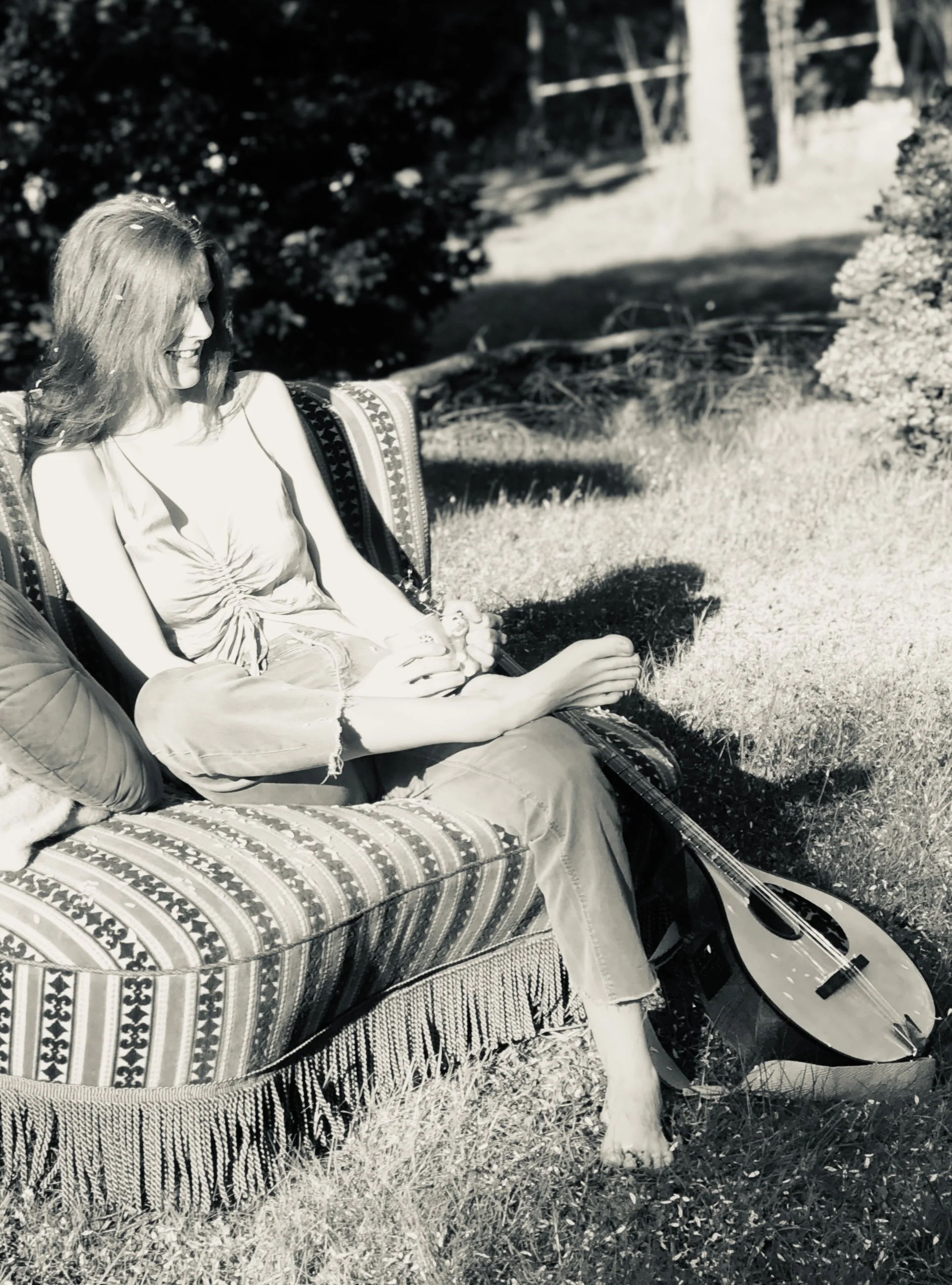 Eine Frau sitzt entspannt auf einem verzierten Gartensofa im Schatten, umgeben von Pflanzen, mit einer Irish Bouzouki auf dem Rasen neben ihr.