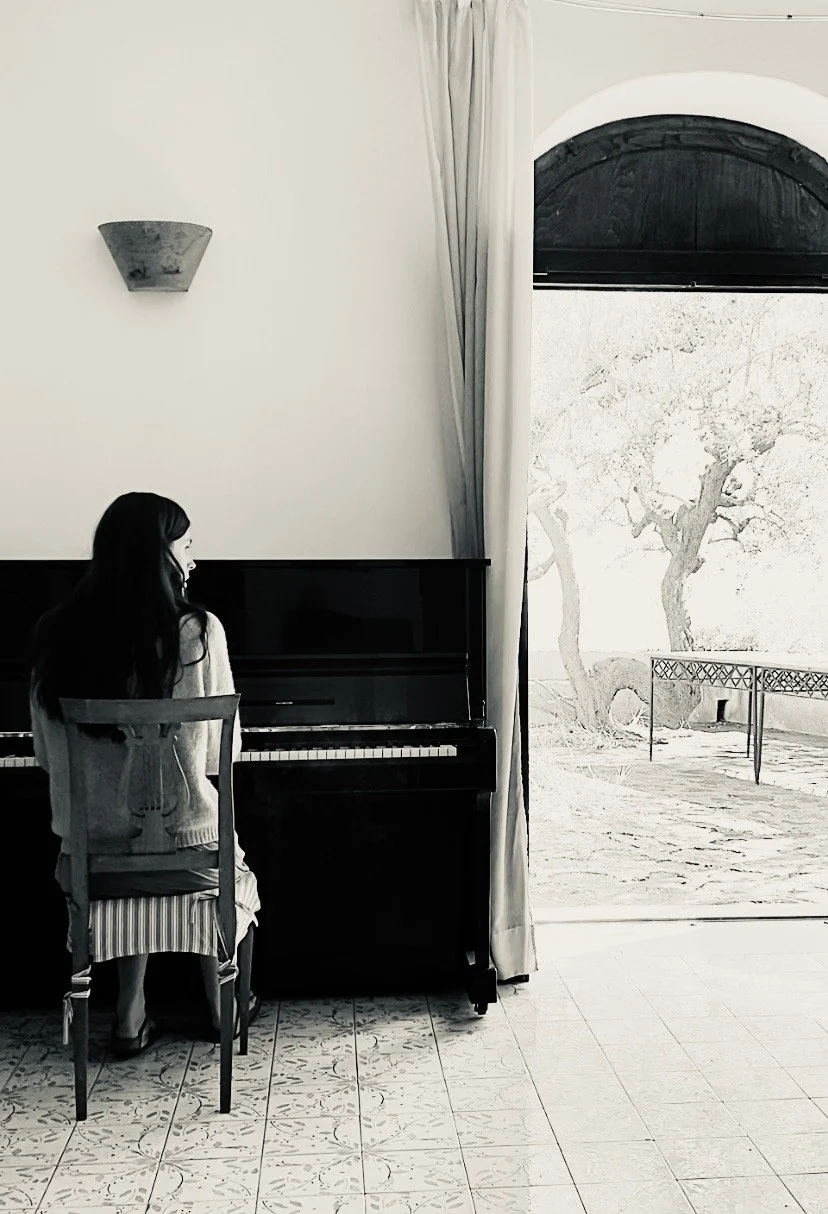 Frau sitzt an einem Klavier im Wohnzimmer, blickt nach draußen auf einen Garten mit Bäumen und einer Terrasse, im monochromen Stil.
