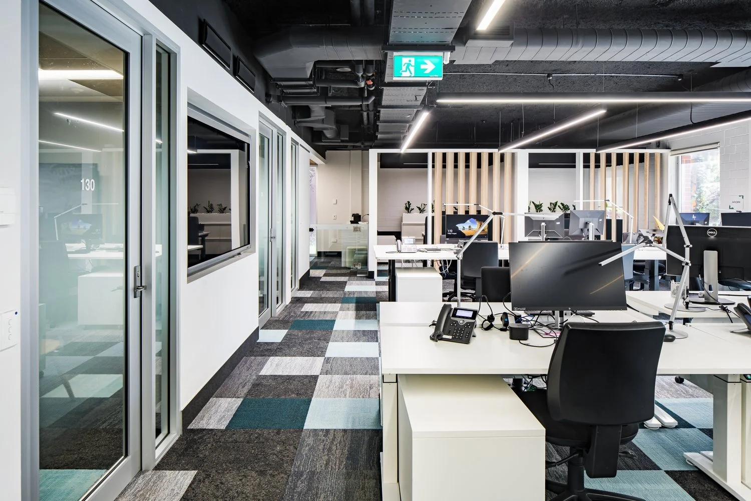 Modern open-plan office with desks, computers, and office equipment; glass-walled meeting rooms on the left; ceiling with exposed ducts and linear lighting; exit sign hanging from ceiling.