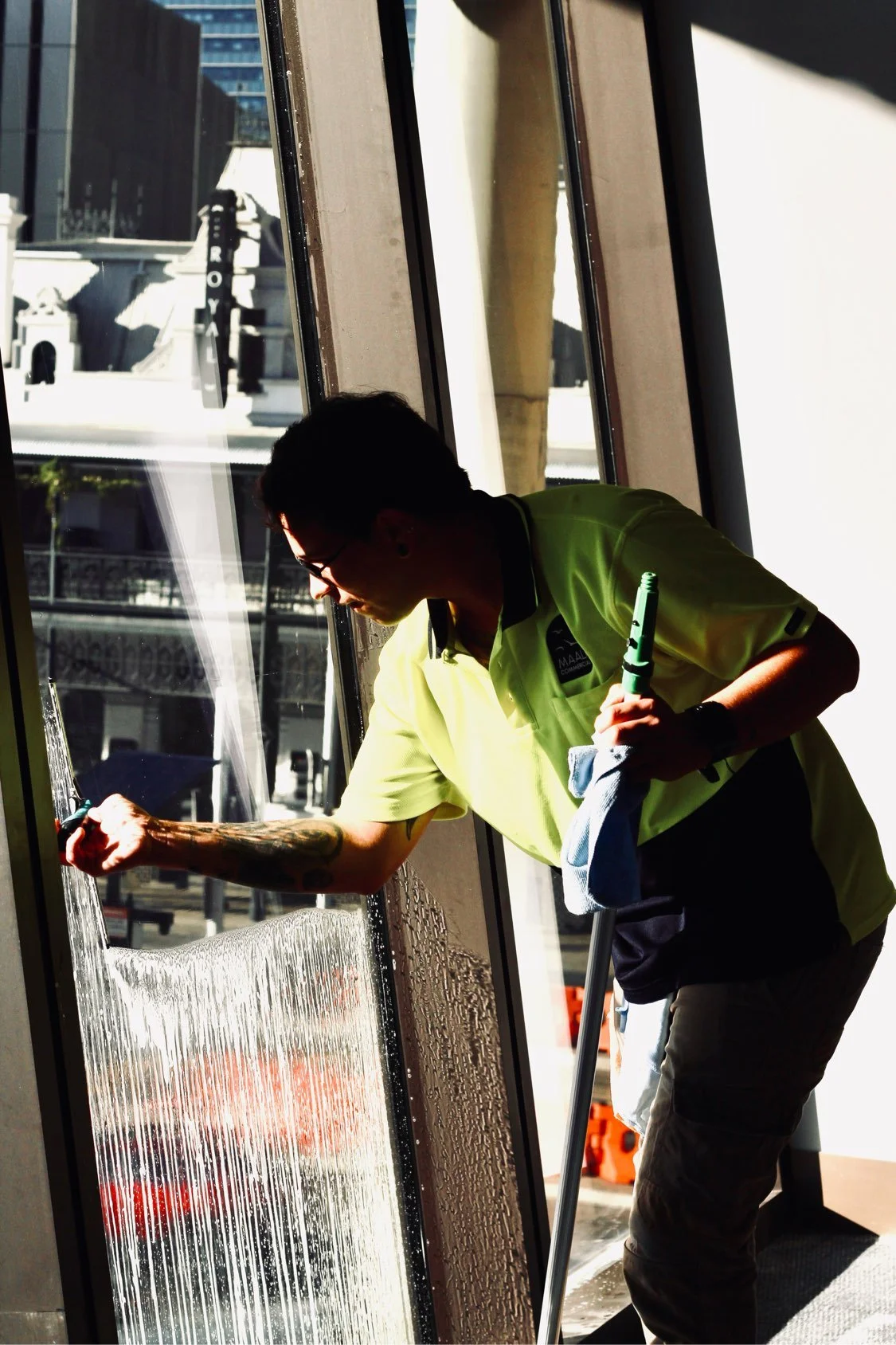 A man cleaning a large glass window on a sunny day, wearing a yellow shirt, glasses, and a watch, with the cityscape visible outside.