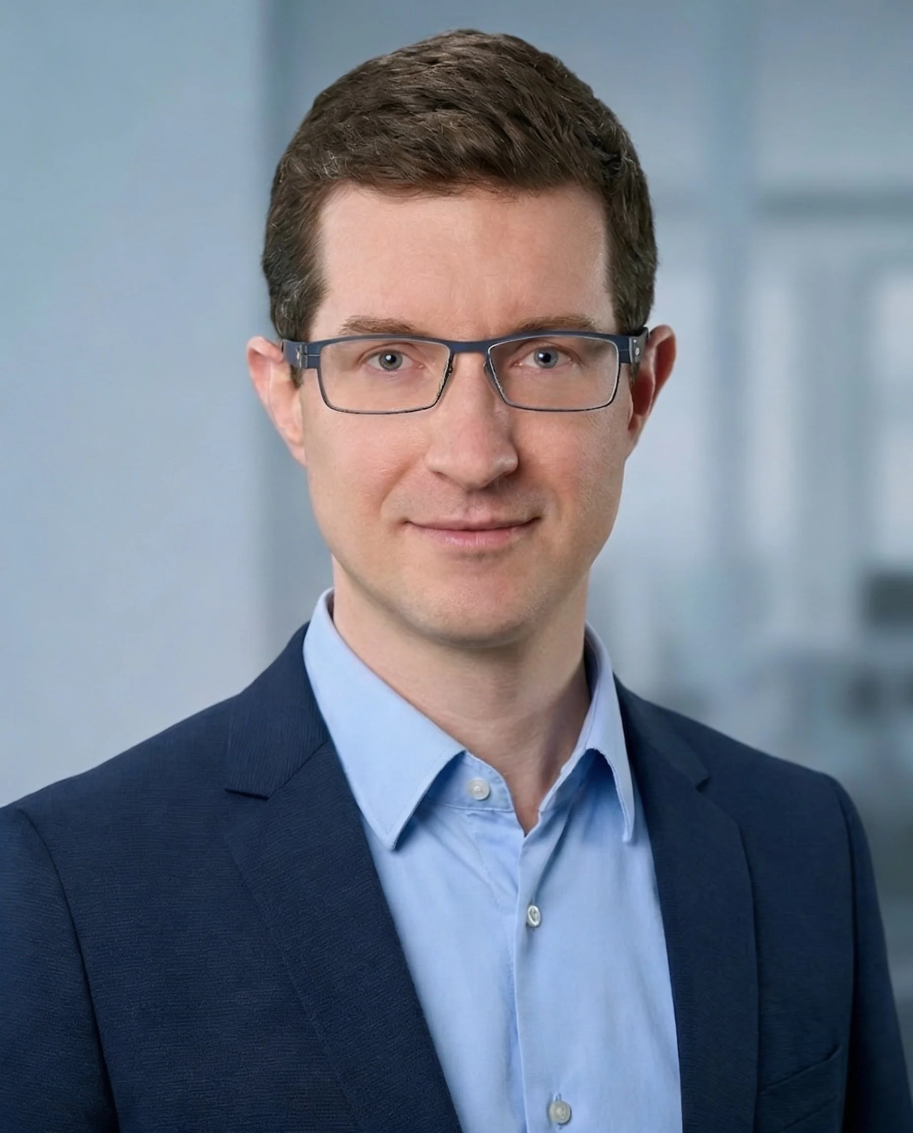 Portrait eines professionellen Coaches für gesundheitsorientierte Führung in dunkelblauem Business‑Sakko und hellblauem Hemd vor modernem Büro‑Hintergrund.