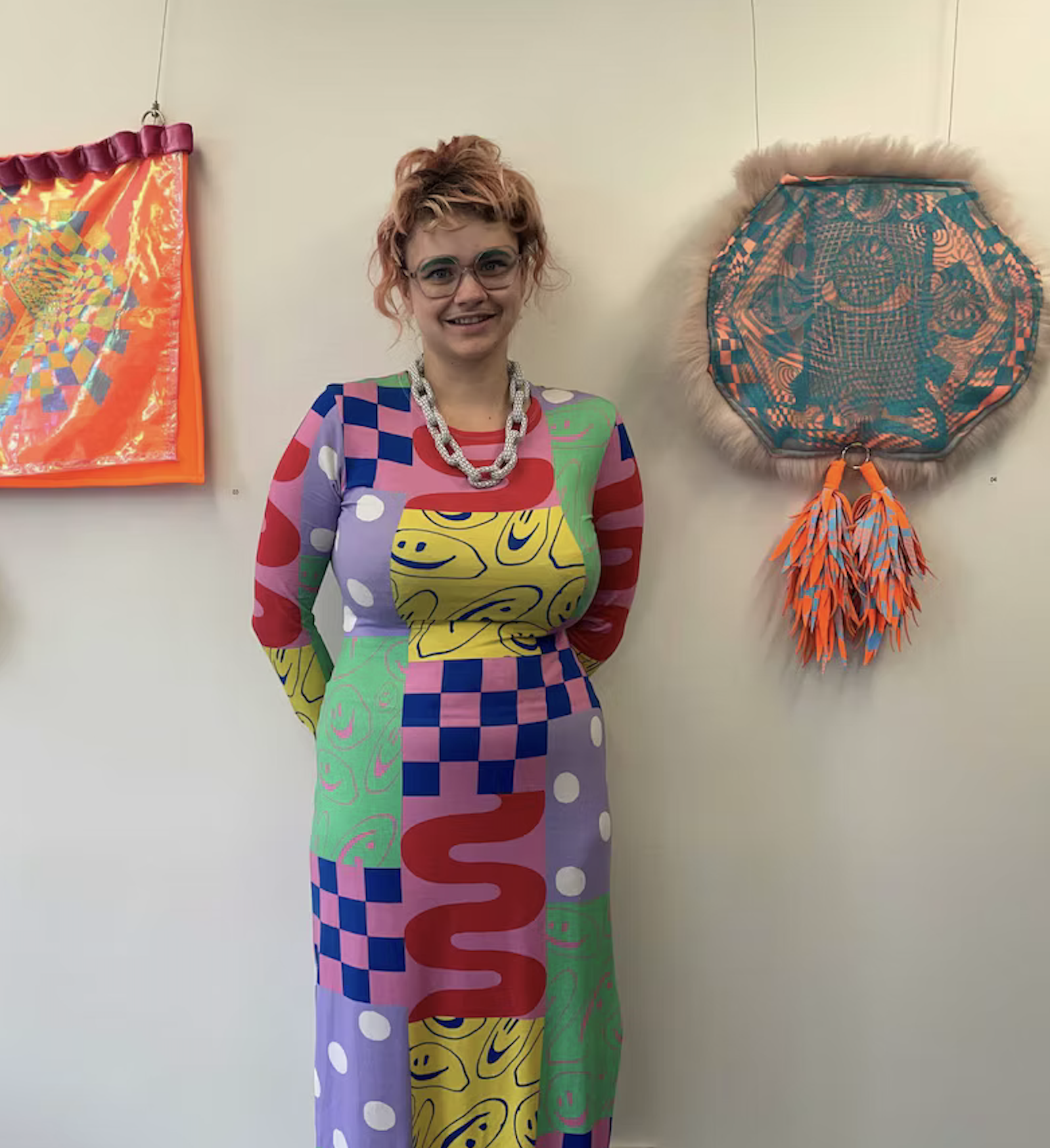 Sandy Sanderson , An artist wearing glasses and a colorful patterned dress with smiley faces, standing in front of a white wall with abstract art pieces that are made from soft sculpture .