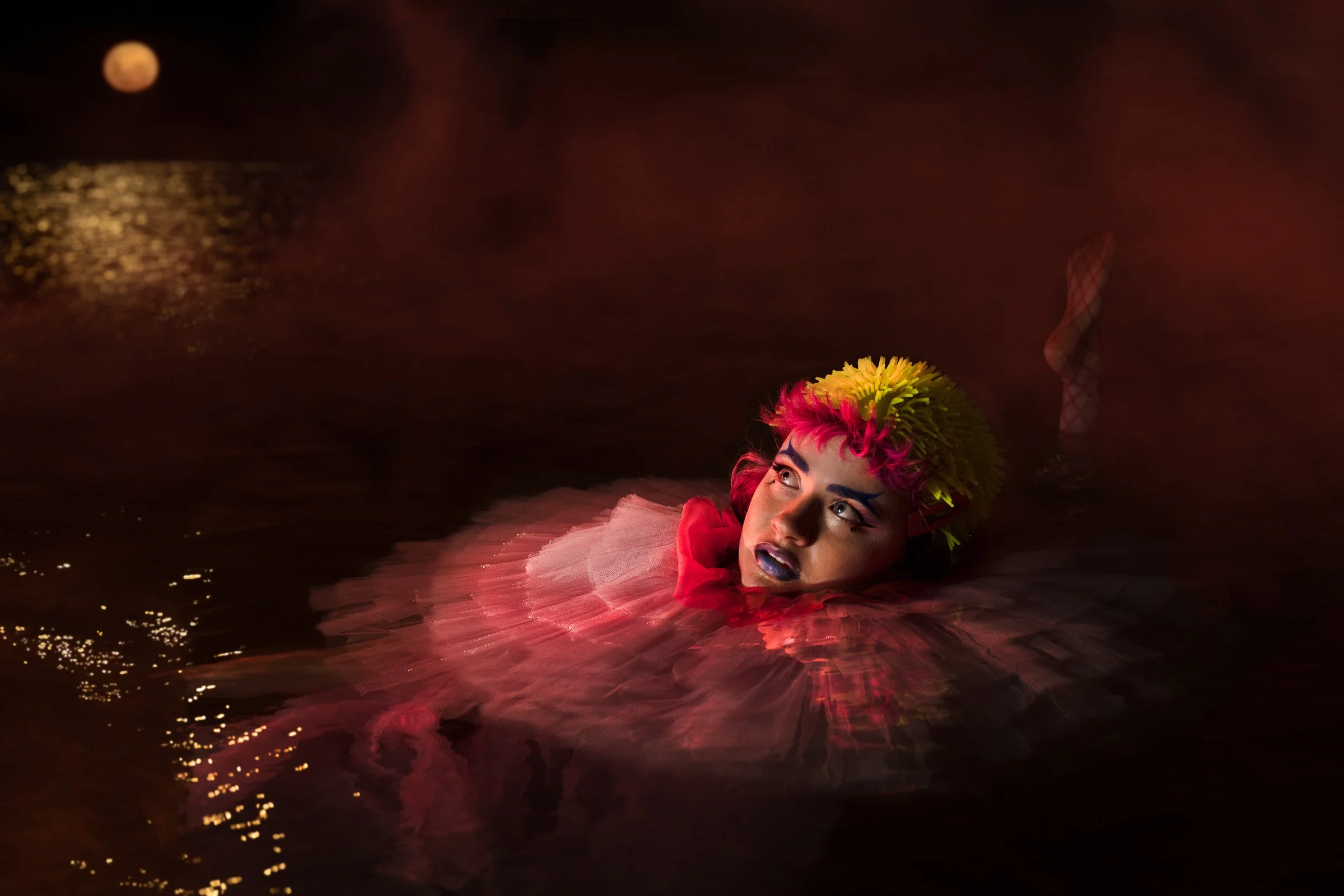 Sandy Sanderson , An artist with colorful hair and dramatic makeup floating in water during a nighttime water show, with the moon and reflections visible in the background. Photo by Edwina Jill Richards for International women's day