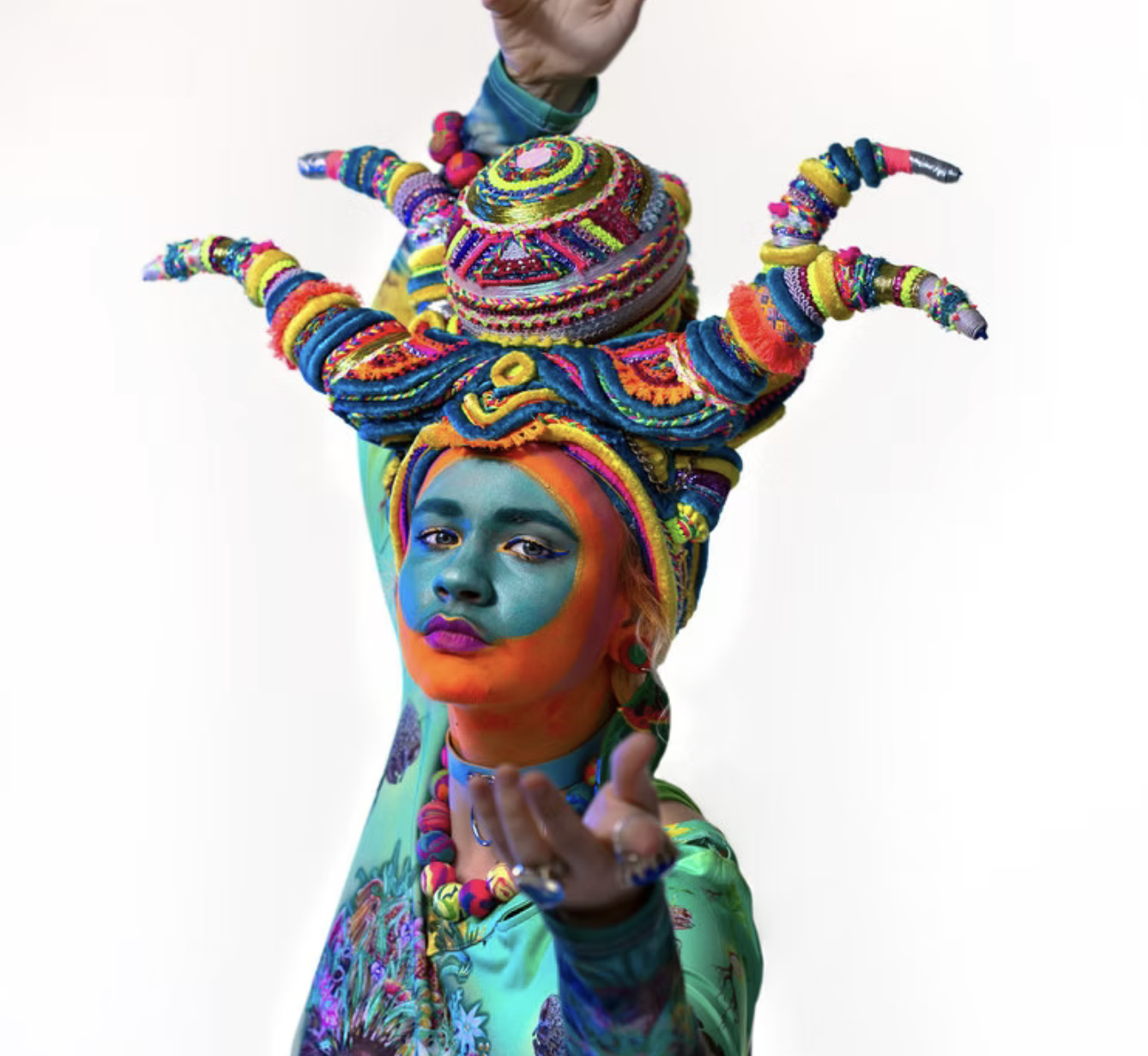 Sandy Sanderson , An artist with vibrant, multicolored face makeup wearing a large headpiece made of numerous coiled and beaded accessories, gesturing toward the camera against a plain white background.
