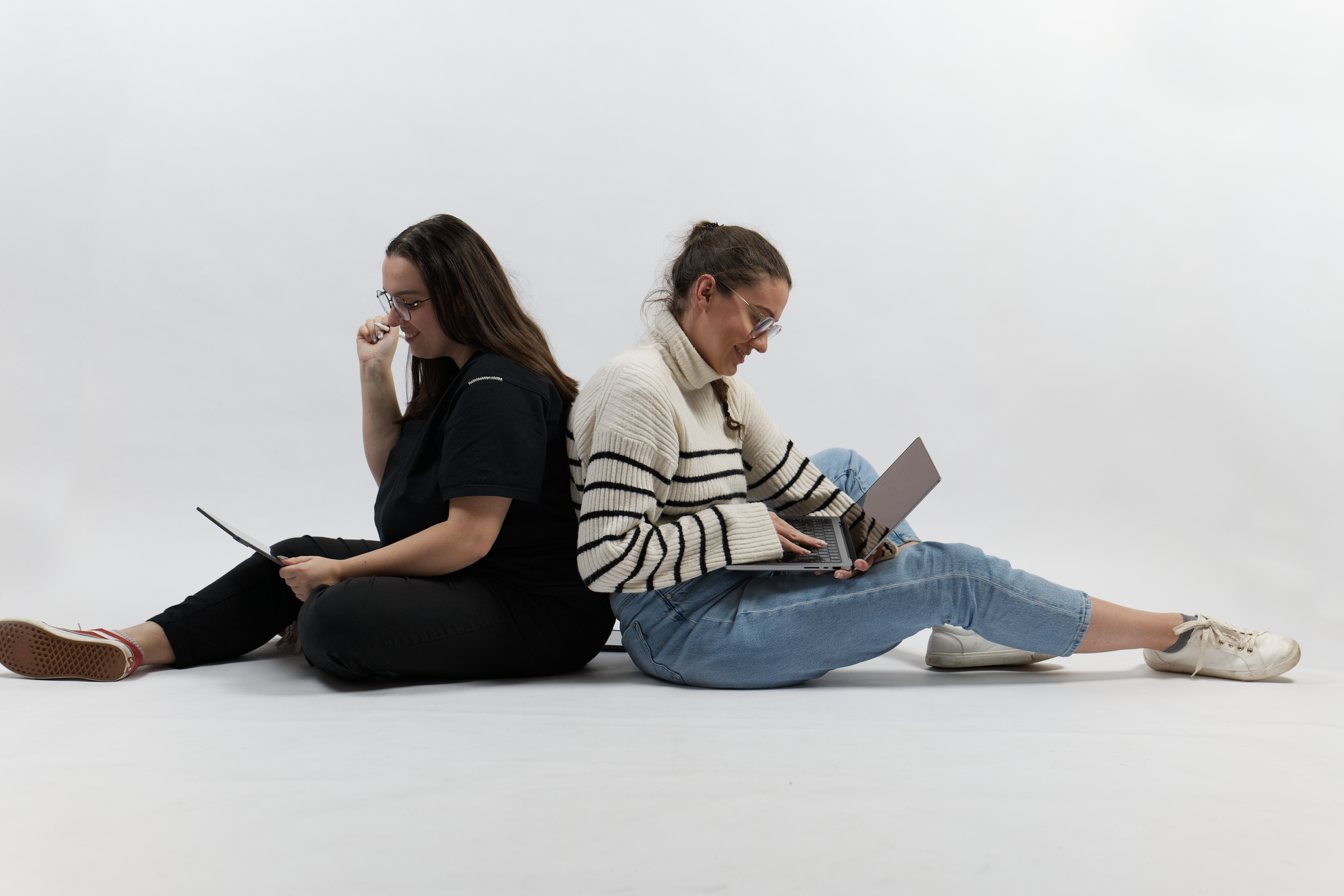 Deux femmes assises dos à dos, utilisant un ordinateur portable et un téléphone portable.