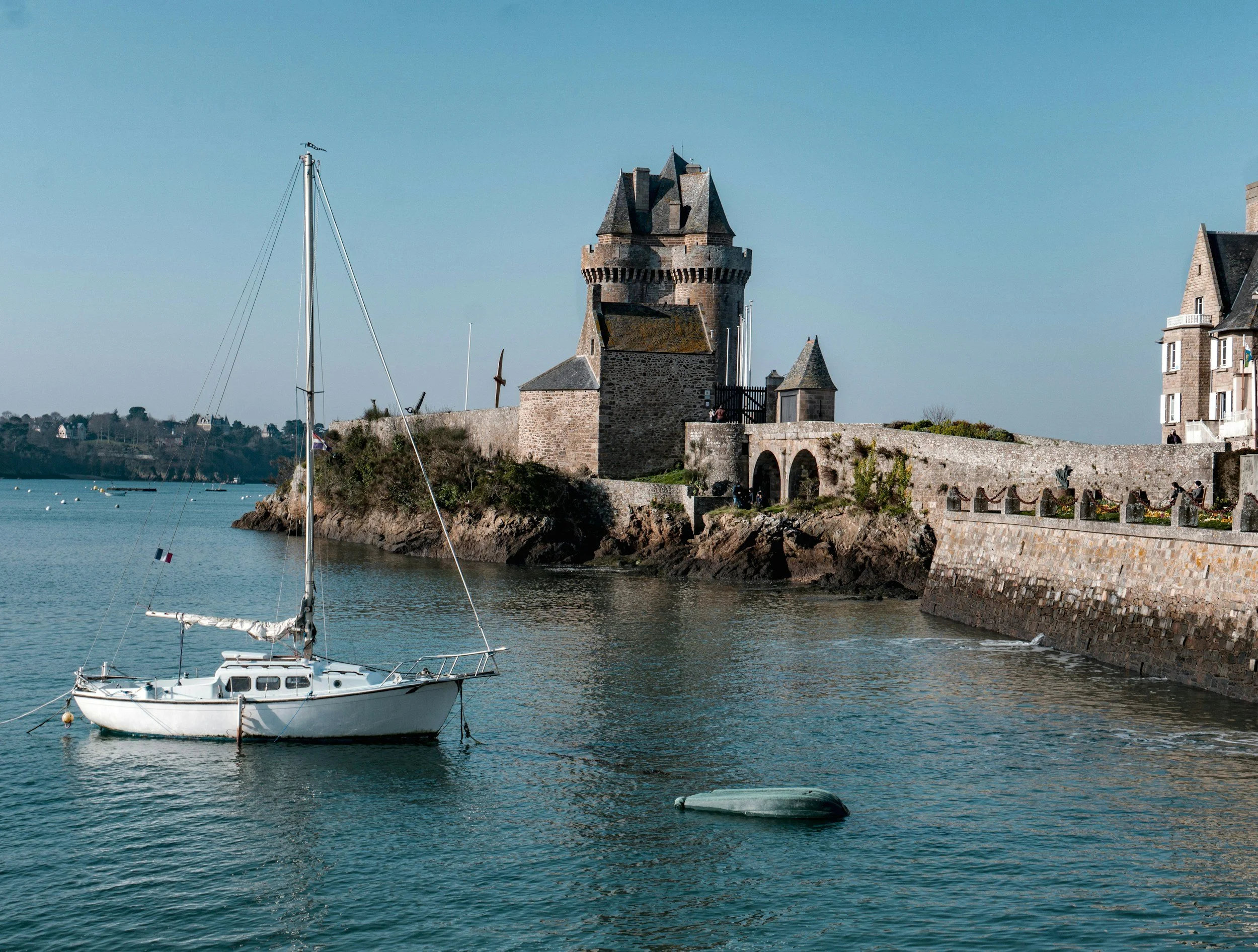 Un bateau de plaisance blanc dans une baie calme avec un château en pierre et une maison en pierre sur la côte dans le fond, sous un ciel clair.