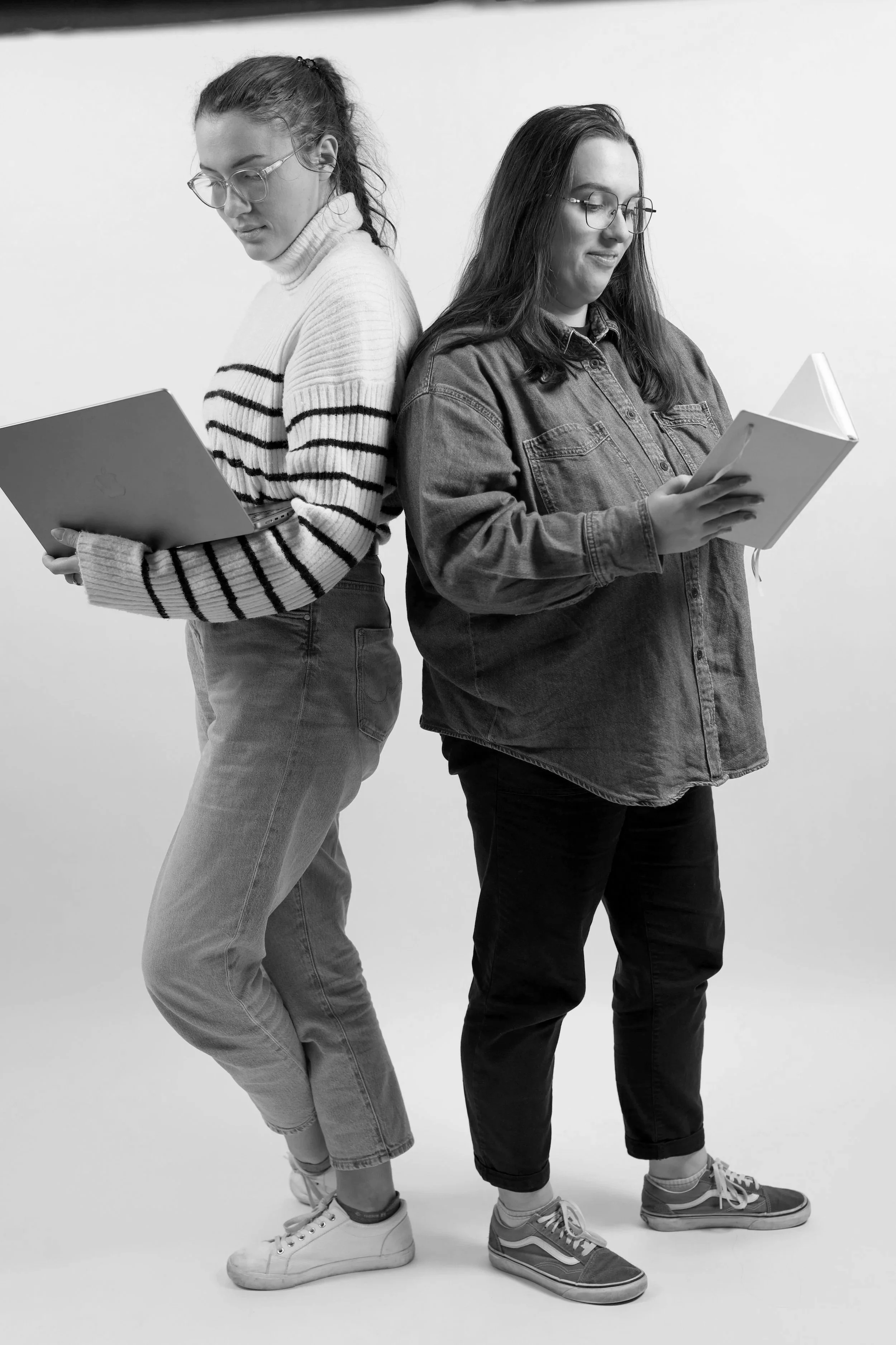 Deux femmes debout dos à dos, lisant des livres. L'une porte un pull à rayures, l'autre une veste en jean.